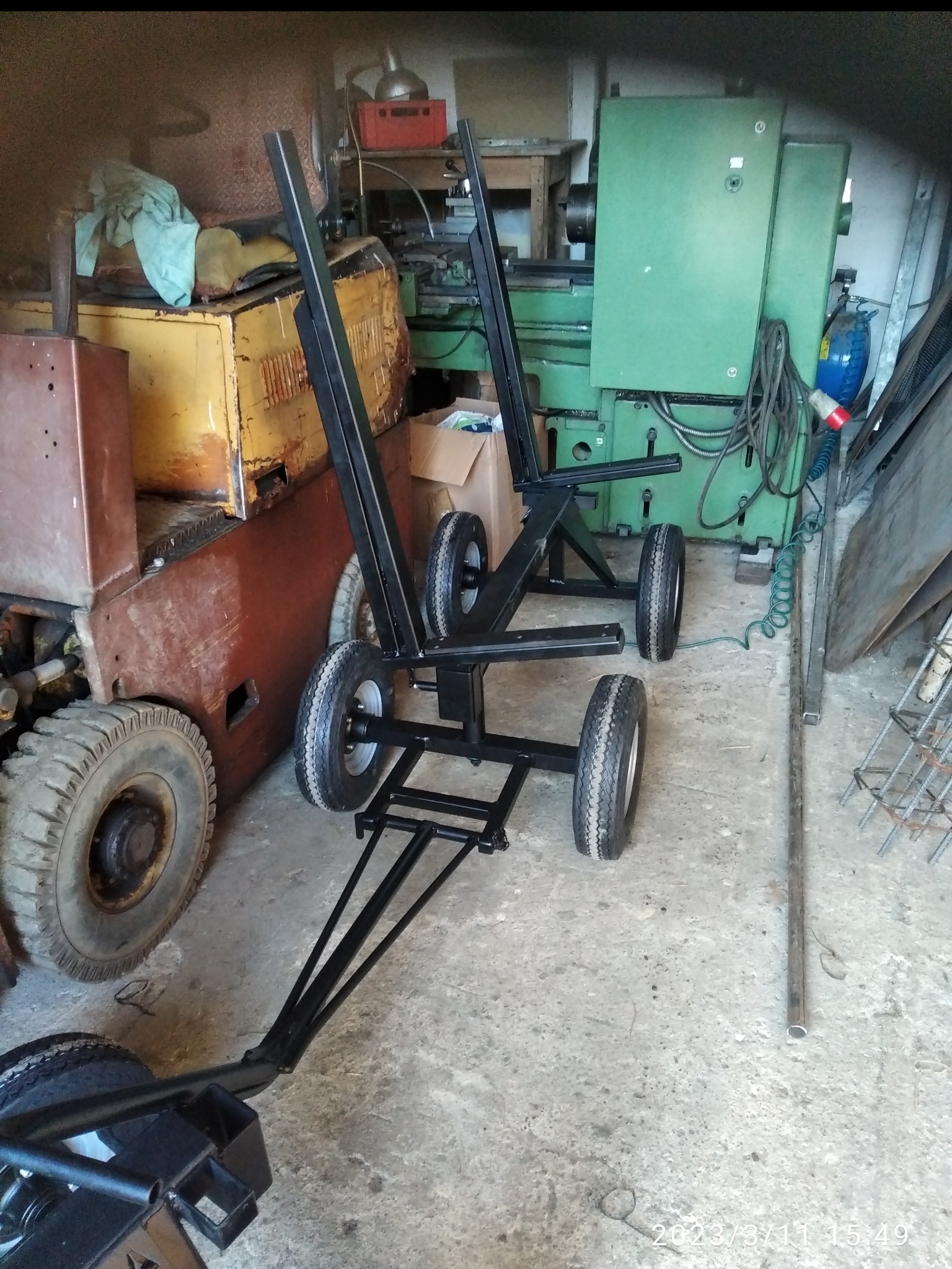 Metalowa konstrukcja na kołach, z pionowymi wspornikami, na tle warsztatu z maszynami. Widoczne elementy konstrukcyjne i spawane łączenia.