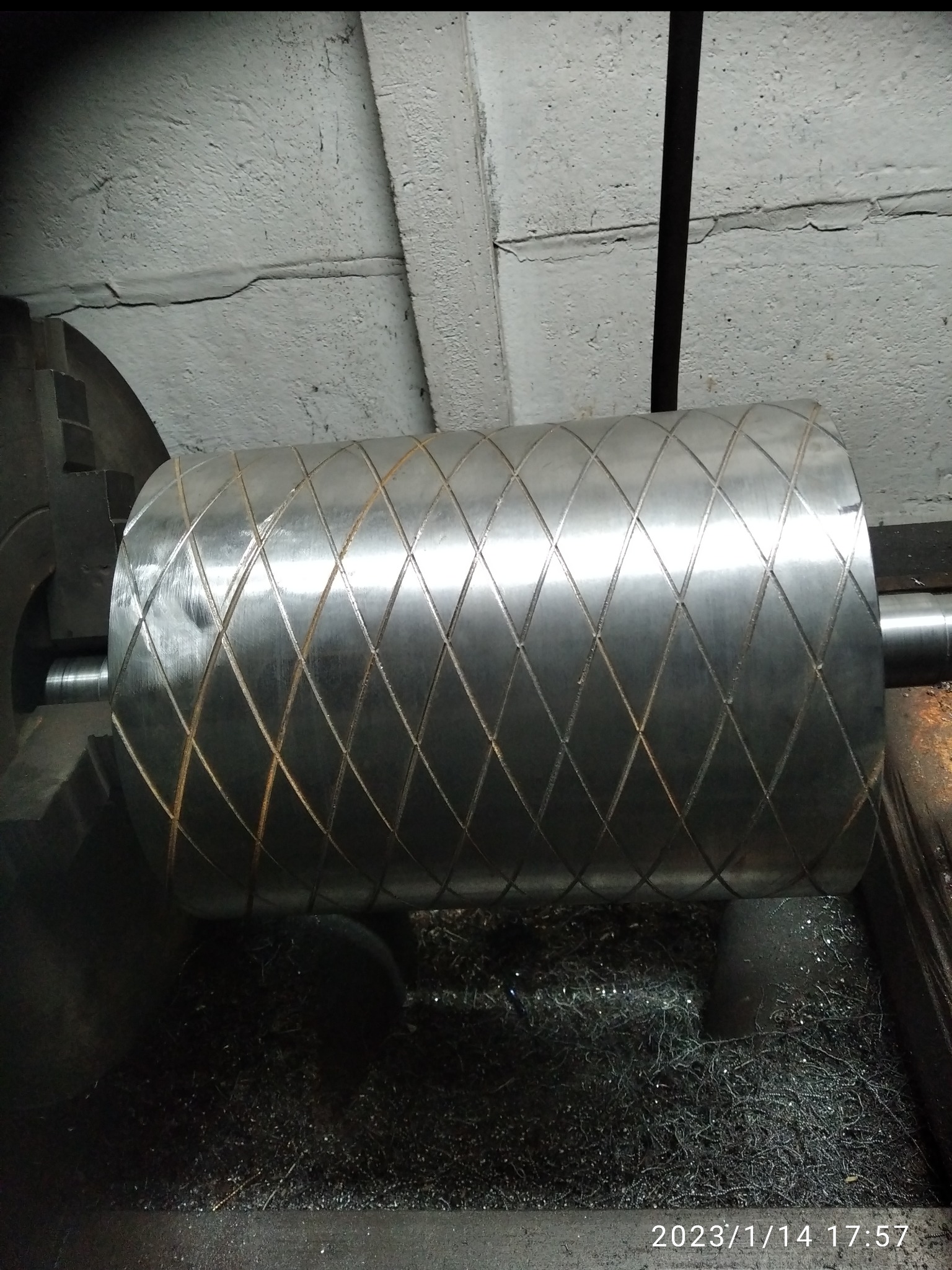 Metalowy wałek z diamentowym wzorem w tokarce, otoczony wiórami. Detal obróbki skrawaniem, widoczne ślady pracy.