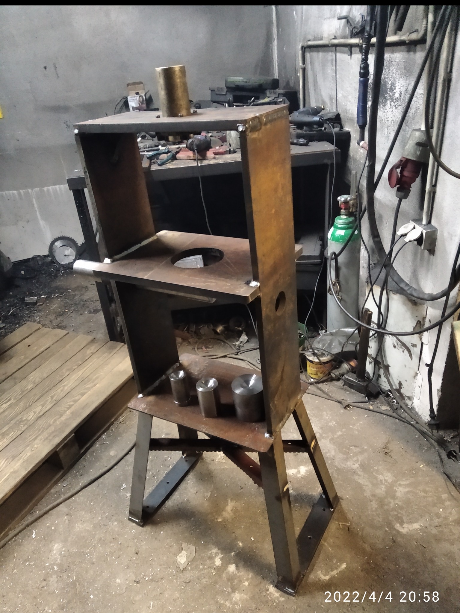 Metalowa konstrukcja spawana z profili, widoczne spawy, otwory i elementy cylindryczne. Surowy warsztat z butlą gazową w tle.