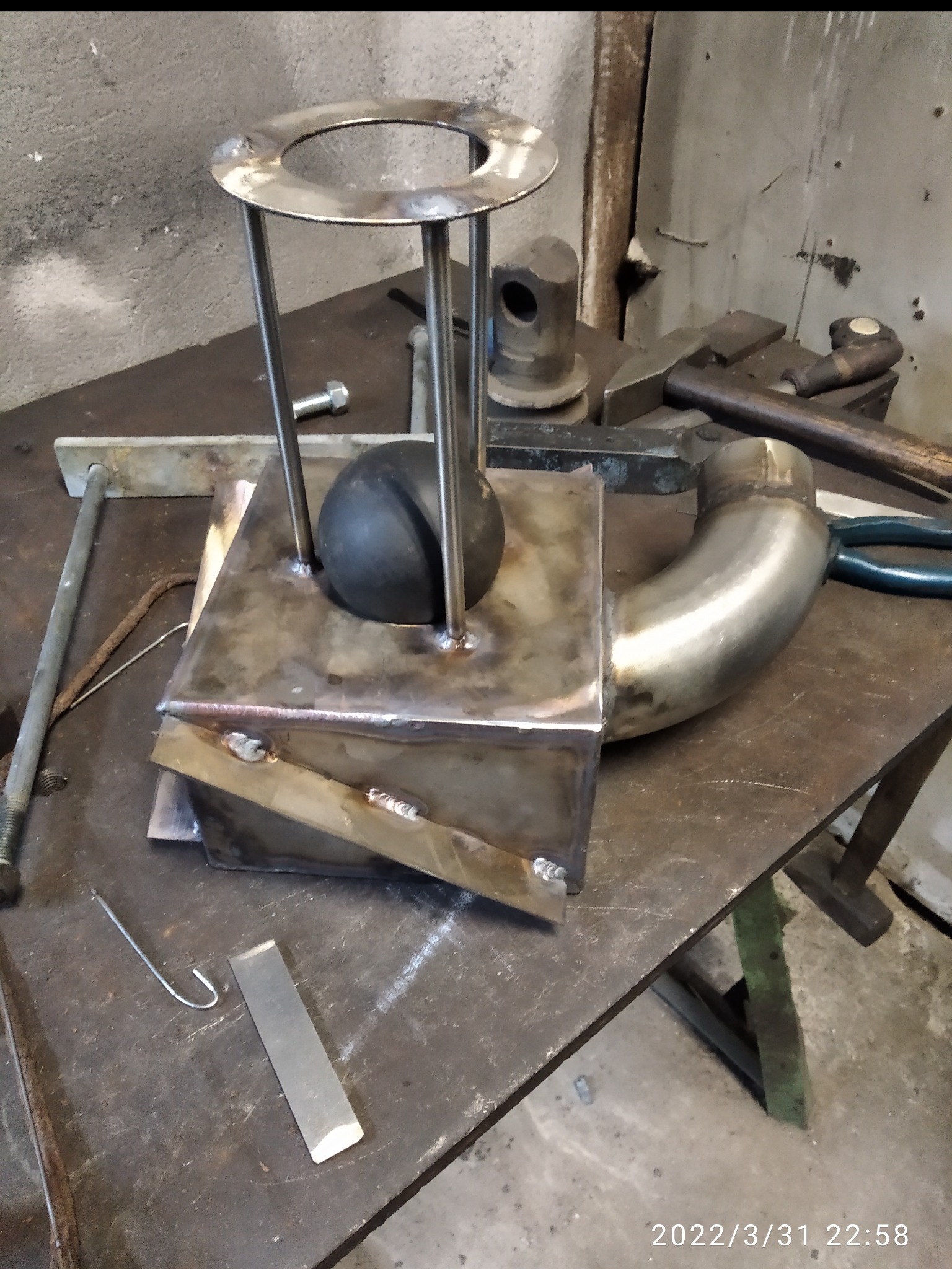 Spawany metalowy element z rurką, kulką i pierścieniem na warsztatowym stole. Widoczne ślady spawania i narzędzia.