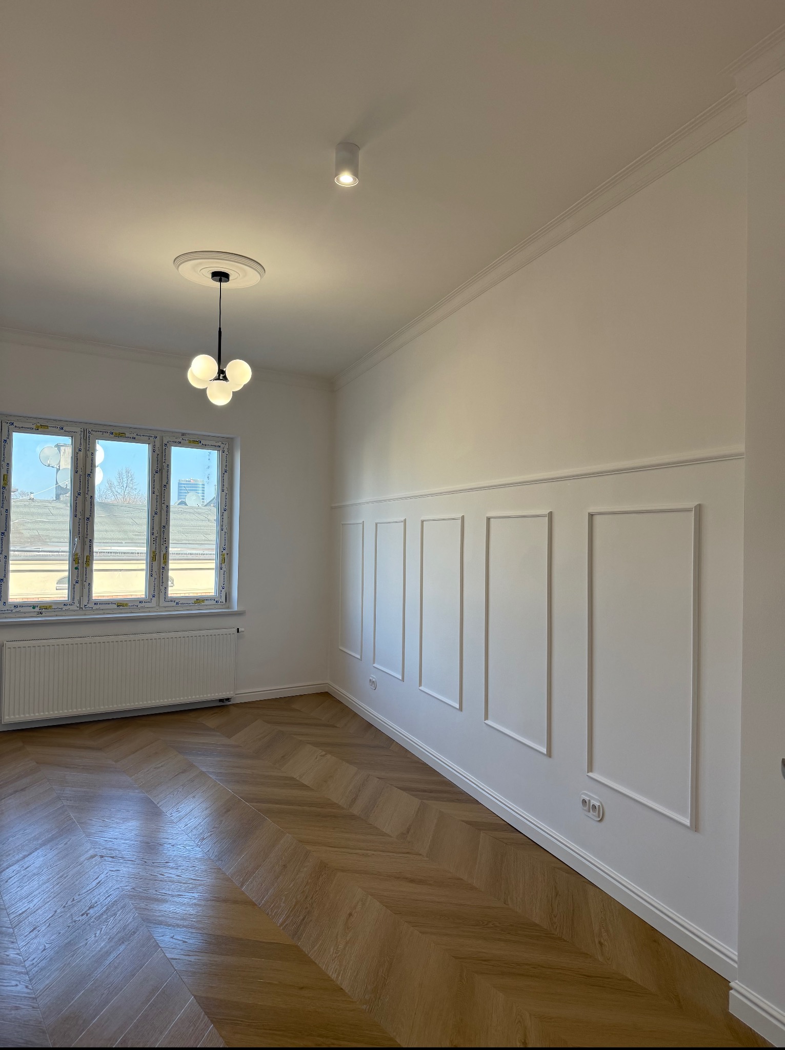 Eleganckie wnętrze po wykończeniu: parkiet w jodełkę, białe ściany z dekoracyjnymi panelami, okno z widokiem na miasto i nowoczesna lampa sufitowa.