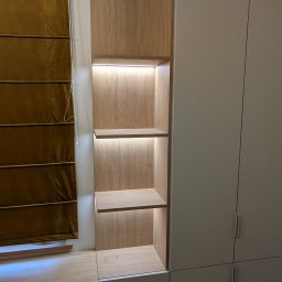 Vihajster - Bartłomiej Jaskuła - Drewniana szafa z półkami podświetlonymi LED, zintegrowana z zabudową, obok okno z brązową roletą rzymską. Jasne, naturalne wykończenie drewna i minimalistyczny design.