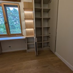 Vihajster - Bartłomiej Jaskuła - Jasna szafa wnękowa z półkami i biurkiem przy oknie, oświetlenie LED, drewniana podłoga. Widok na zewnątrz przez okno.