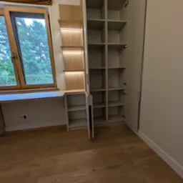 Jasna szafa wnękowa z półkami i biurkiem przy oknie, oświetlenie LED, drewniana podłoga. Widok na zewnątrz przez okno.