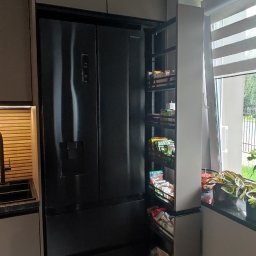 Vihajster - Bartłomiej Jaskuła - Nowoczesna kuchnia z zabudową w odcieniach szarości, czarna lodówka side-by-side i wysuwana szafka cargo obok okna z żaluzjami. Minimalistyczny design.