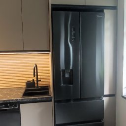 Vihajster - Bartłomiej Jaskuła - Nowoczesna kuchnia z zabudową w odcieniach szarości, czarnym zlewem i lodówką side-by-side Hisense. Uwagę zwraca oświetlenie LED nad blatem i minimalistyczny design.