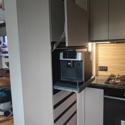 Vihajster - Bartłomiej Jaskuła - Szara zabudowa kuchenna z wysuwanym blatem i ekspresem do kawy. Górna szafka uchylona, widoczne szuflady z czarnymi uchwytami i fragment płyty grzewczej.