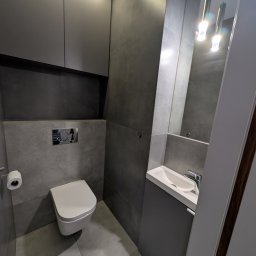 Vihajster - Bartłomiej Jaskuła - Nowoczesna, szara łazienka z podwieszaną toaletą, umywalką z szafką i lustrem z oświetleniem. Minimalistyczny design, płytki imitujące beton, eleganckie wykończenie.