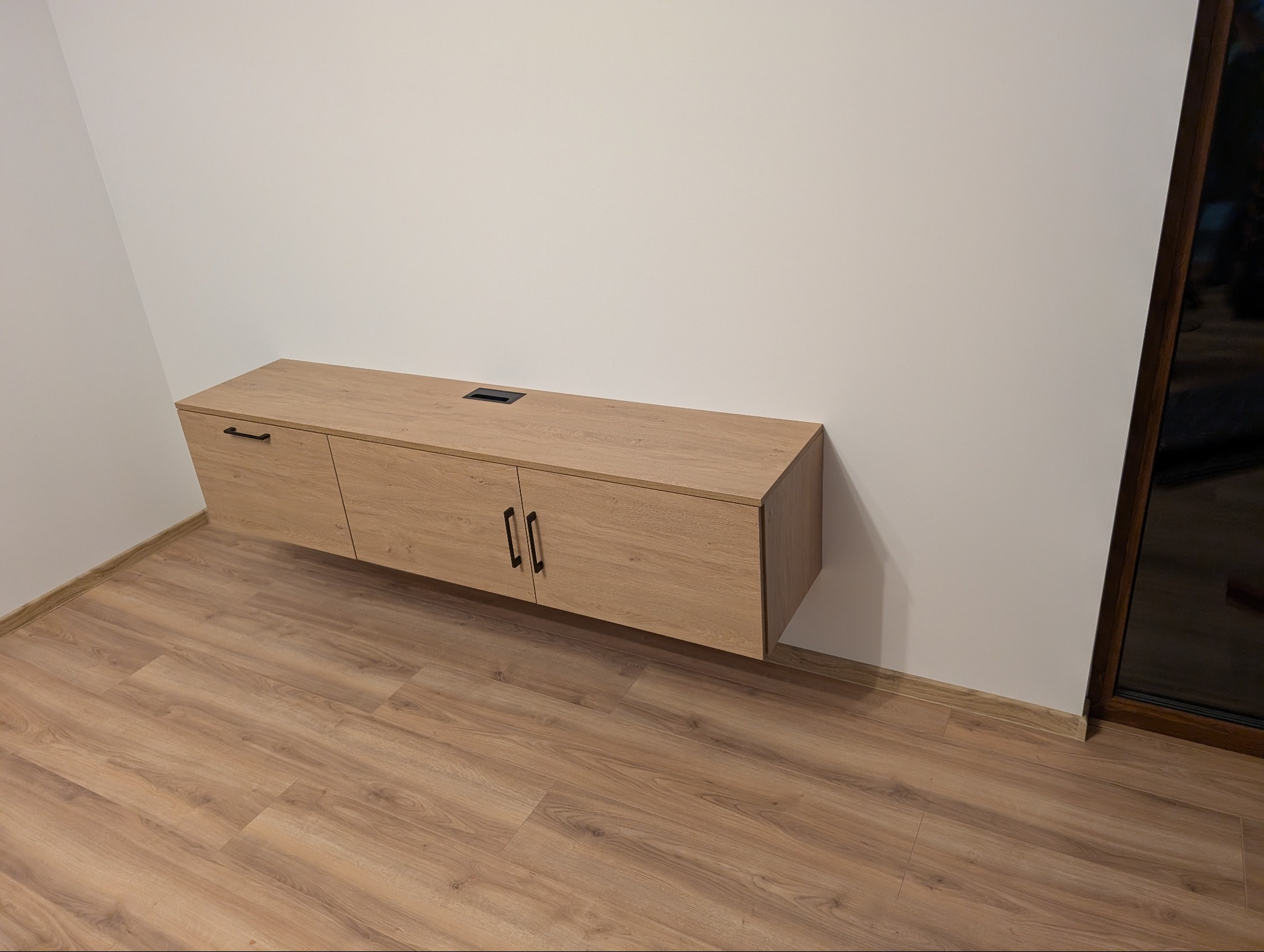 Podwieszana szafka RTV z blatem imitującym drewno, z czarnymi uchwytami, zamontowana na białej ścianie nad panelami podłogowymi. Widoczny otwór na kable.