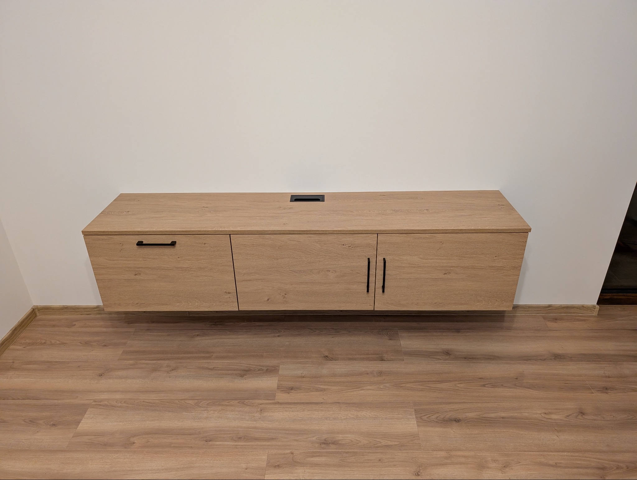 Wisząca szafka RTV z jasnego drewna z czarnymi uchwytami, zamontowana na białej ścianie nad panelami podłogowymi. Widoczny otwór na kable na górze szafki.