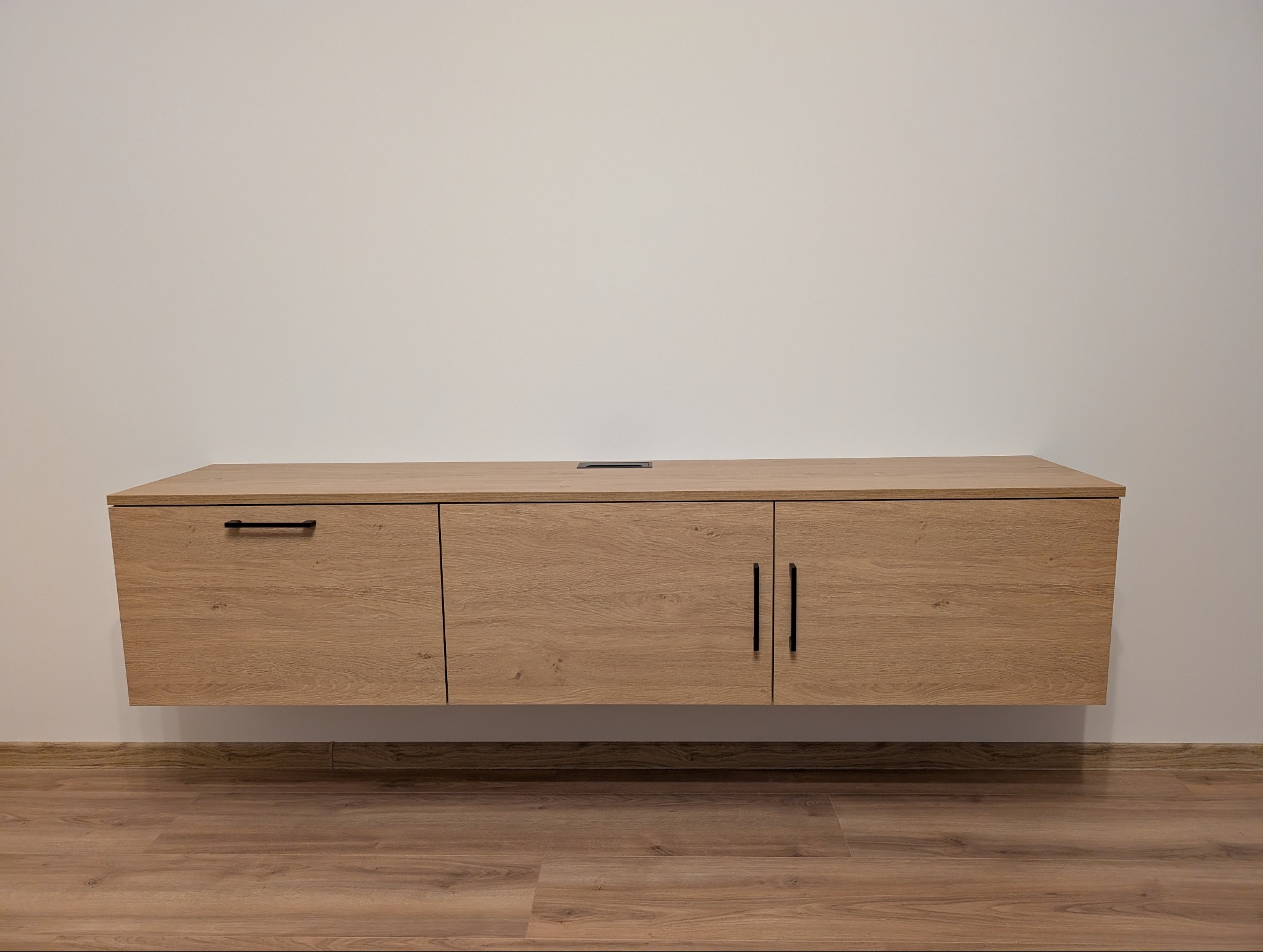 Jasna, drewnopodobna szafka RTV wisząca na ścianie, z czarnymi uchwytami, na tle białej ściany i podłogi z paneli. Widoczny otwór na kable.