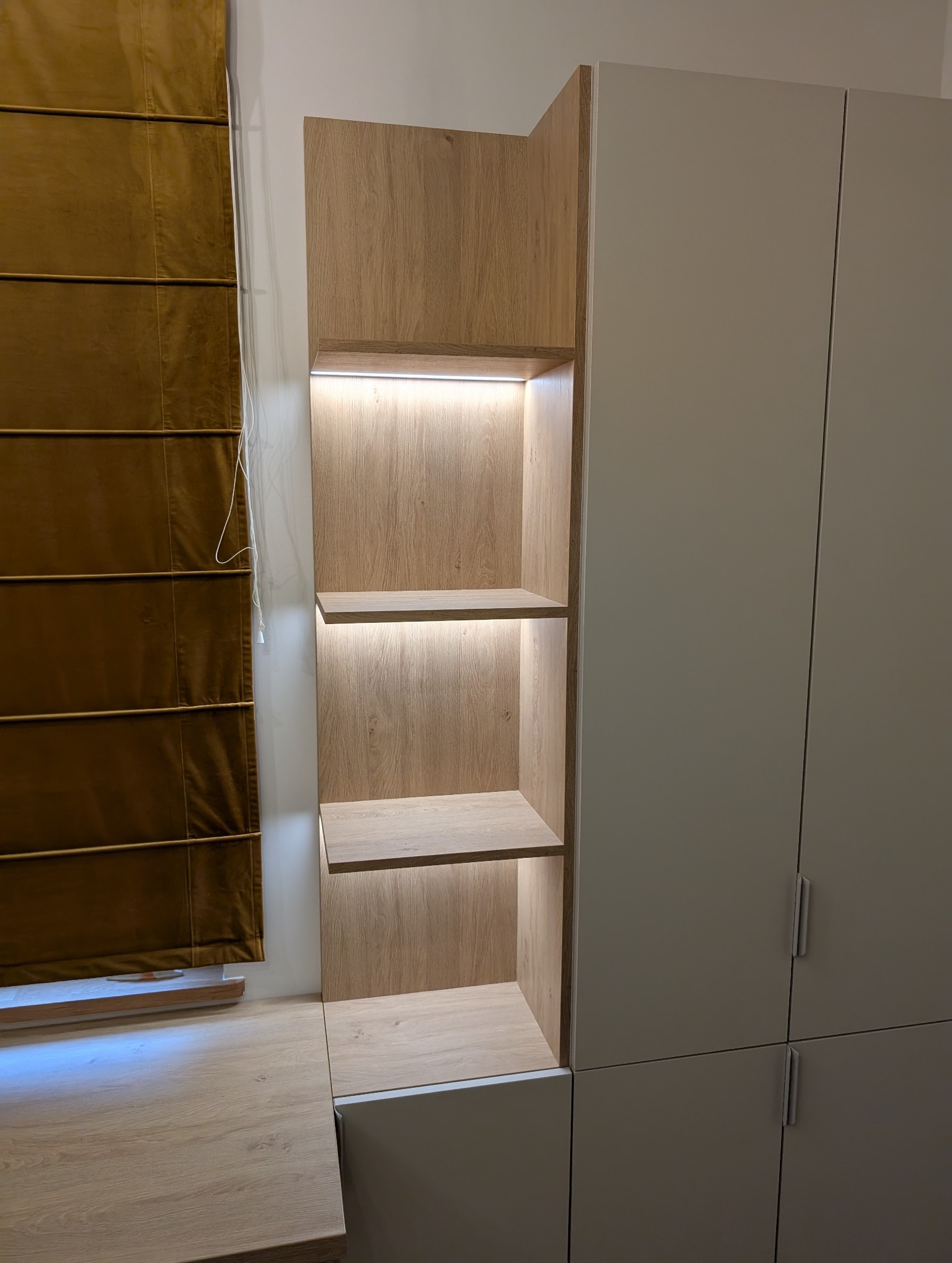 Drewniana szafa z półkami podświetlonymi LED, zintegrowana z zabudową, obok okno z brązową roletą rzymską. Jasne, naturalne wykończenie drewna i minimalistyczny design.