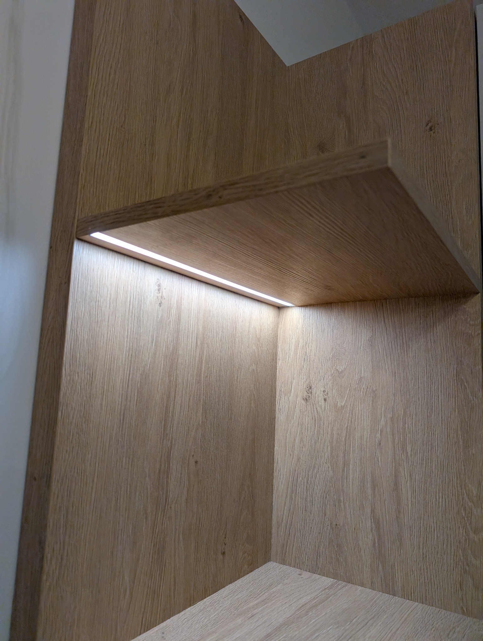 Półka z oświetleniem LED wewnątrz szafy o jasnym, drewnopodobnym wykończeniu. Minimalistyczny design, ciepłe światło podkreśla strukturę drewna.