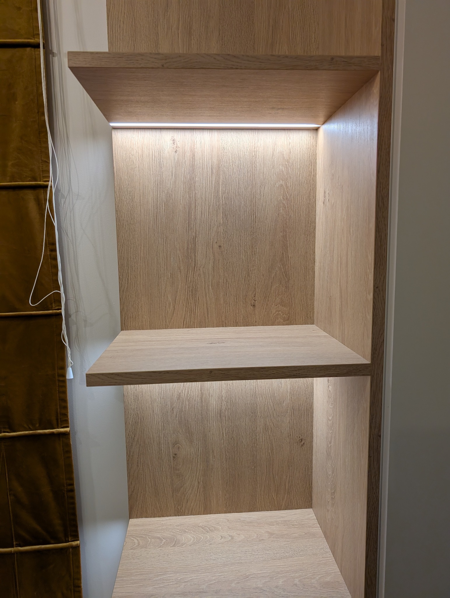 Półki z jasnego drewna z subtelnym oświetleniem LED, tworzące minimalistyczny i funkcjonalny element wystroju wnętrza. Widoczny fragment zasłony w tle.