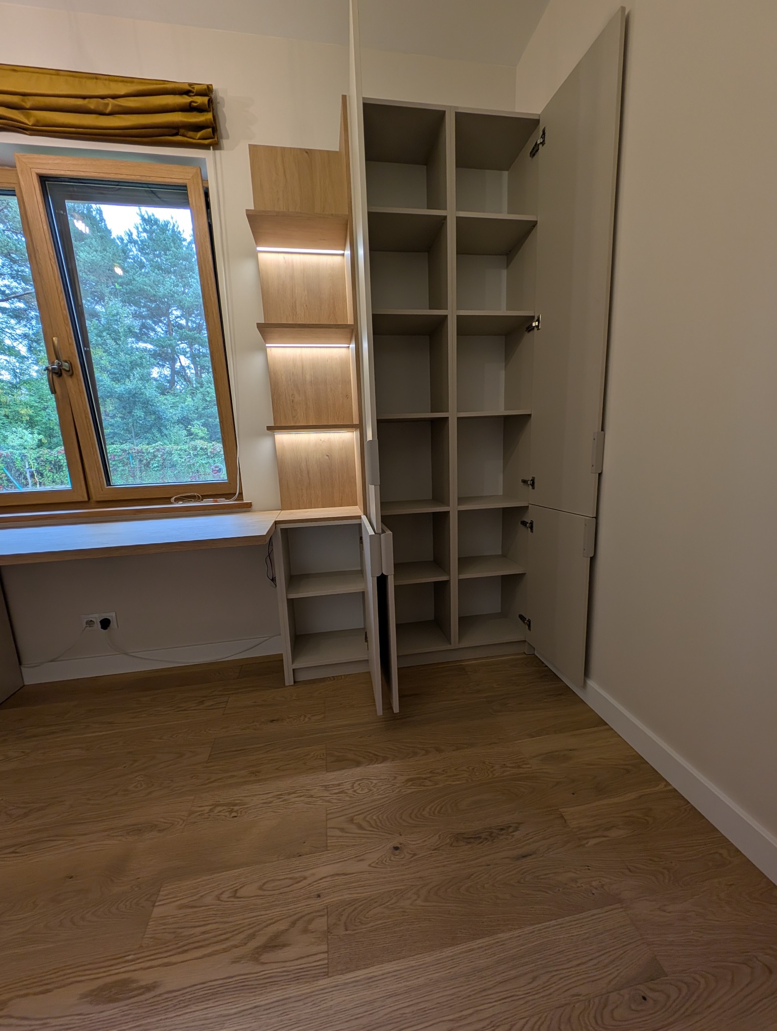 Jasna szafa wnękowa z półkami i biurkiem przy oknie, oświetlenie LED, drewniana podłoga. Widok na zewnątrz przez okno.