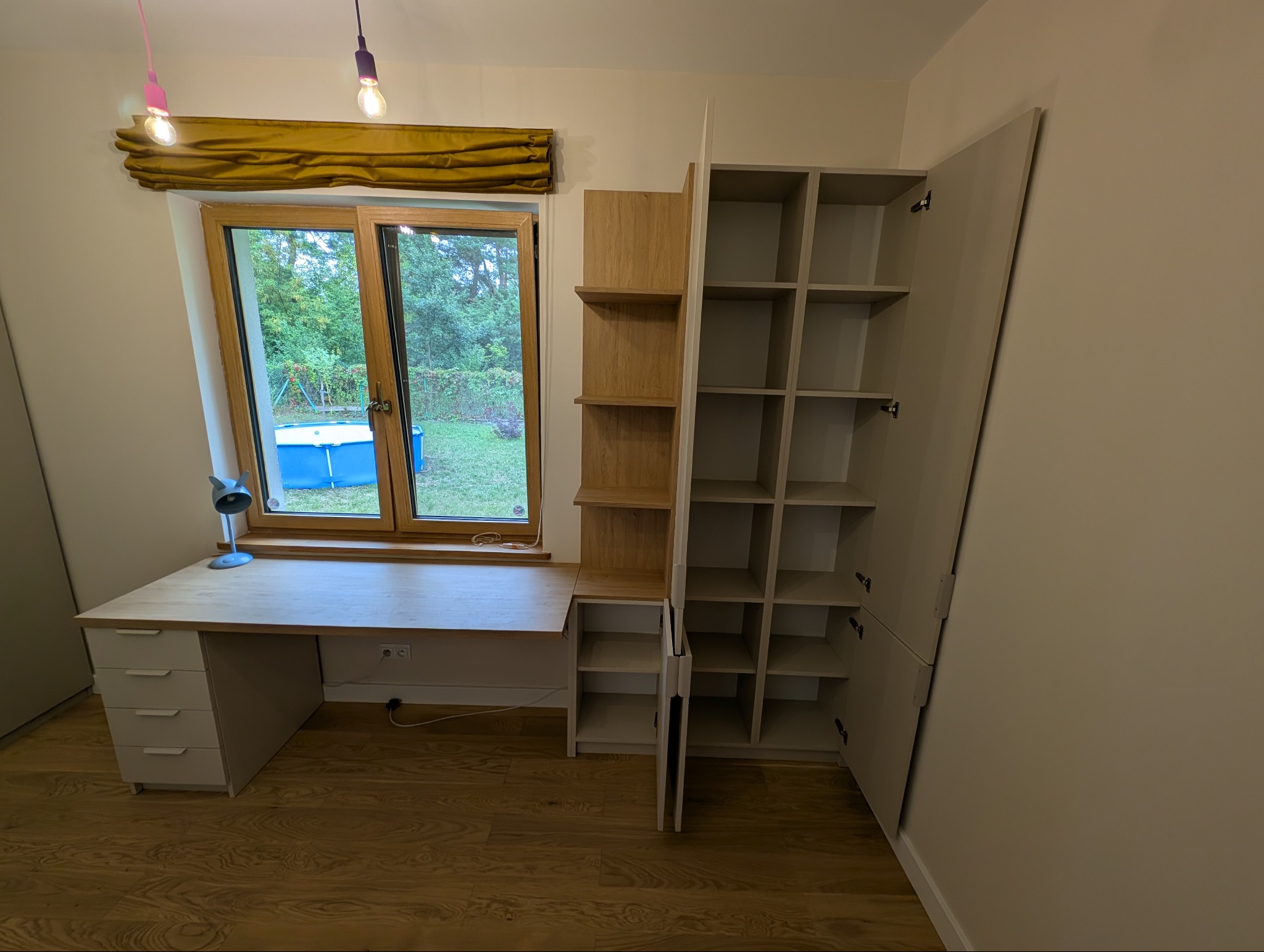 Jasna zabudowa meblowa z biurkiem przy oknie, widoczne półki i szafki. Na zewnątrz widok na ogród z basenem. Całość w nowoczesnym stylu.