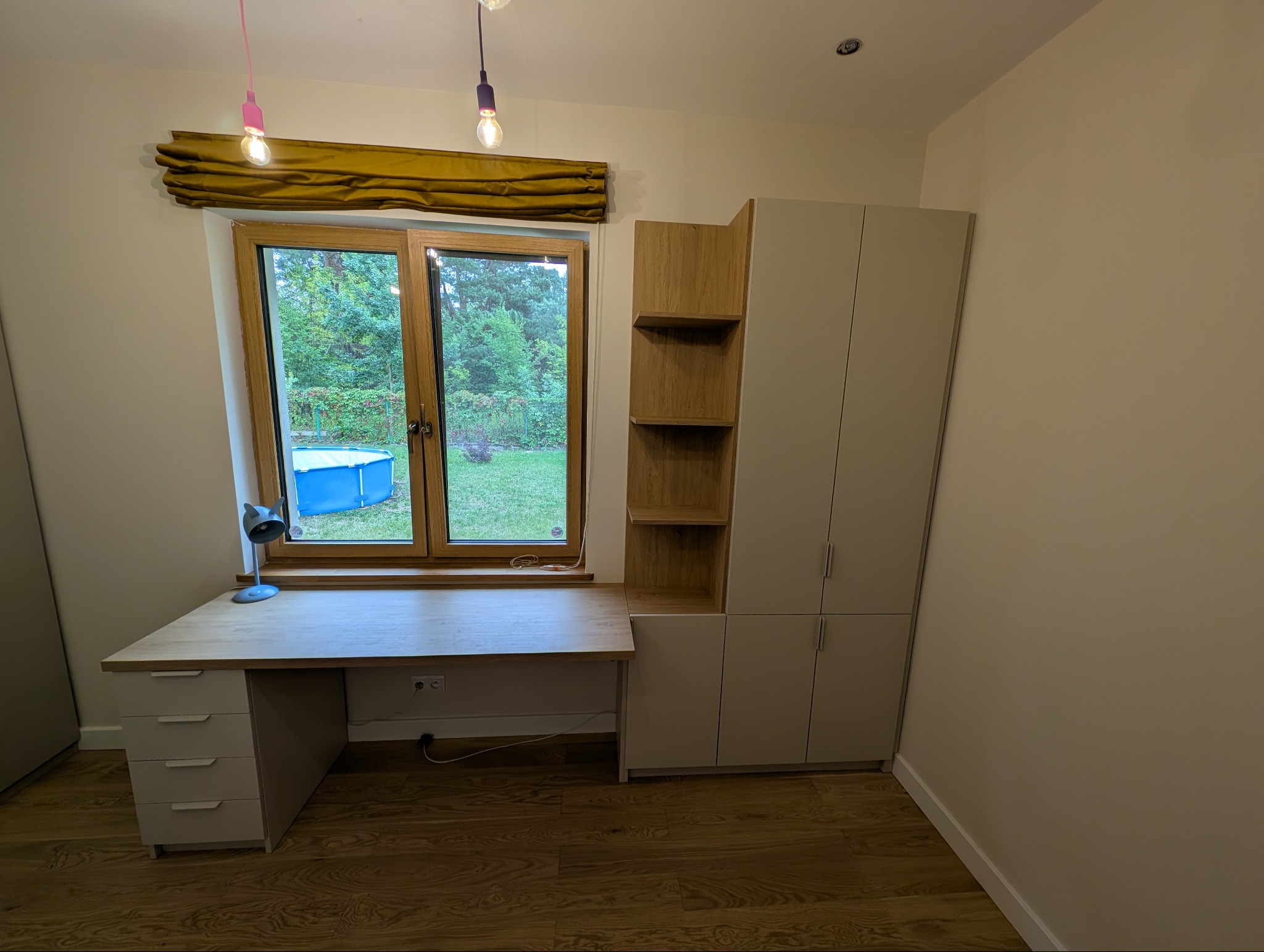 Jasne biurko z szufladami i szafa z półkami w pokoju, widok na ogród przez okno z żółtą roletą. Drewniana podłoga i lampki wiszące.