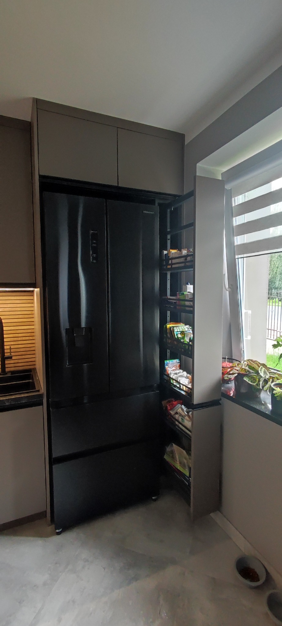 Nowoczesna kuchnia z zabudową w odcieniach szarości, czarna lodówka side-by-side i wysuwana szafka cargo obok okna z żaluzjami. Minimalistyczny design.