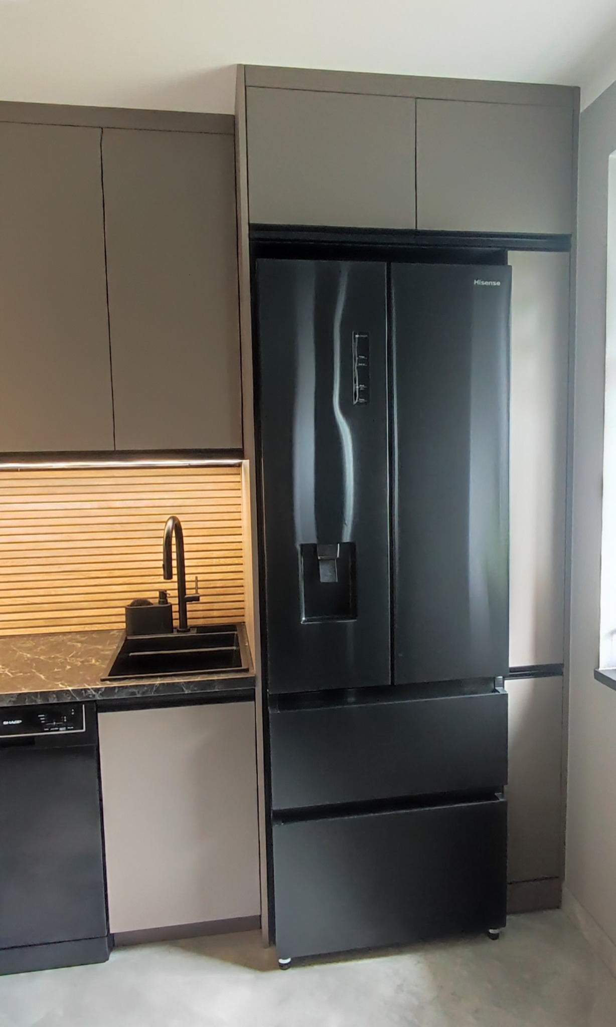 Nowoczesna kuchnia z zabudową w odcieniach szarości, czarnym zlewem i lodówką side-by-side Hisense. Uwagę zwraca oświetlenie LED nad blatem i minimalistyczny design.