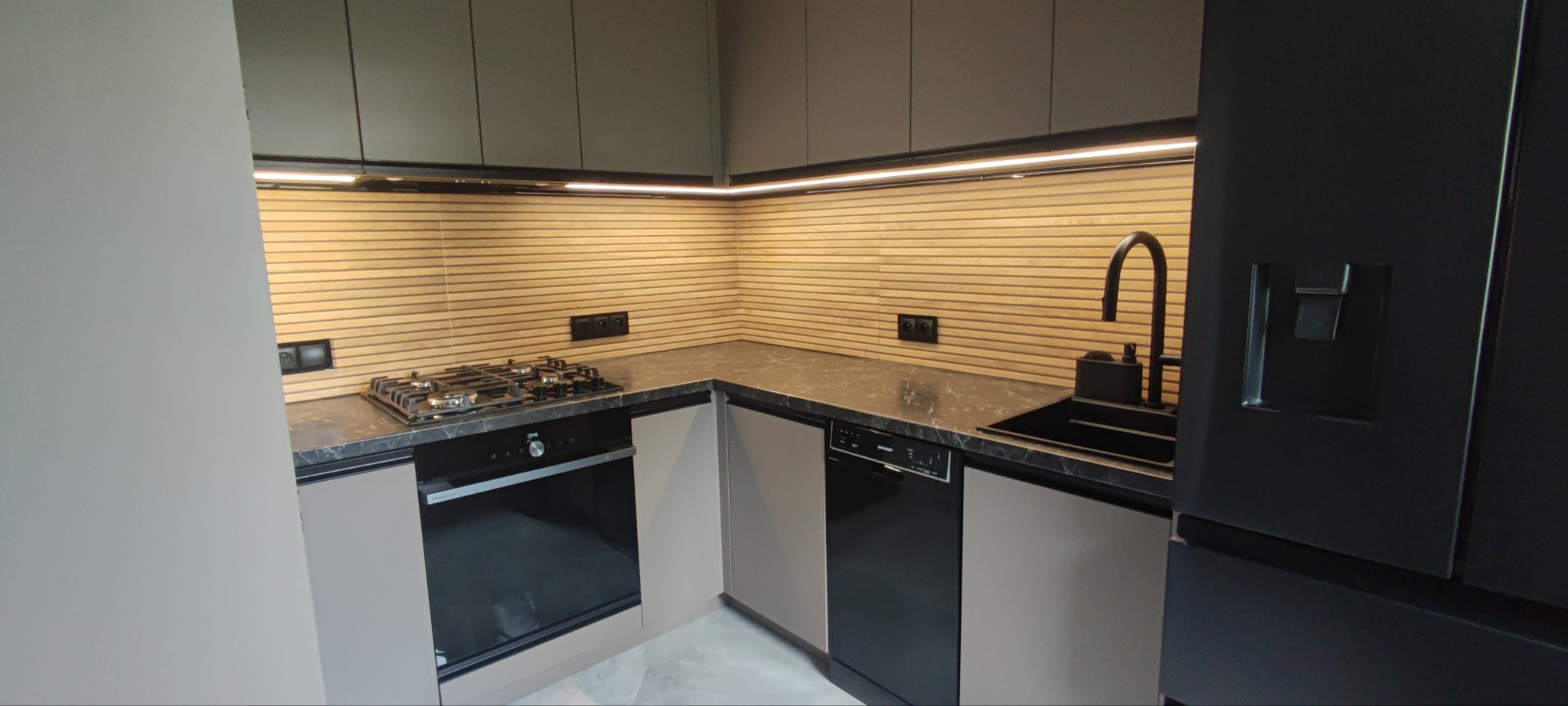 Nowoczesna kuchnia z szarymi szafkami, drewnianym panelem ściennym i czarnymi akcentami: zlew, bateria, lodówka. Oświetlenie LED podkreśla unikalny design.