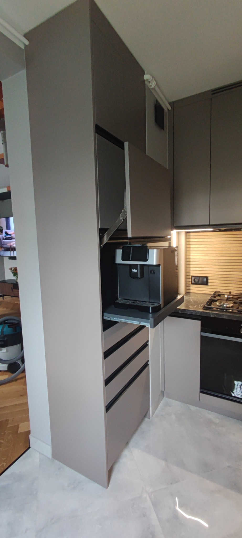 Szara zabudowa kuchenna z wysuwanym blatem i ekspresem do kawy. Górna szafka uchylona, widoczne szuflady z czarnymi uchwytami i fragment płyty grzewczej.