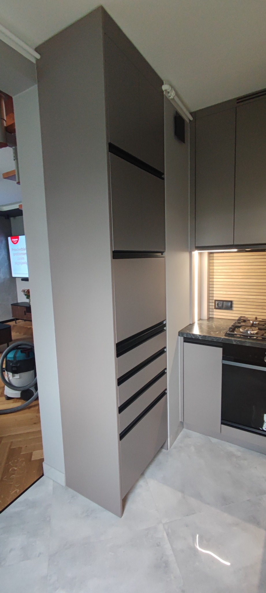 Szara zabudowa kuchenna z szafkami i szufladami z czarnymi uchwytami, oświetlenie LED pod szafkami, widok na płytę grzewczą i piekarnik. Nowoczesny design.