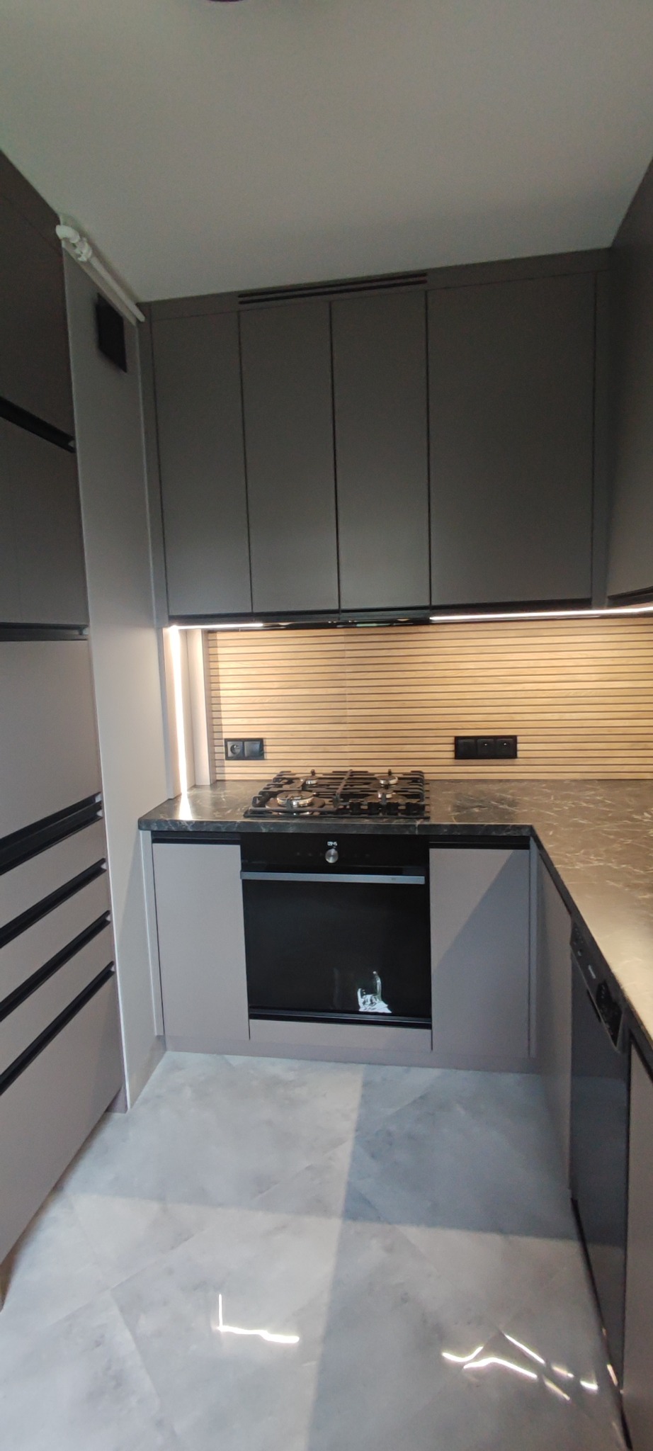 Nowoczesna kuchnia w odcieniach szarości z marmurowym blatem i drewnianą ścianą nad blatem. Oświetlenie LED pod szafkami górnymi. Widok na płytę gazową i piekarnik.