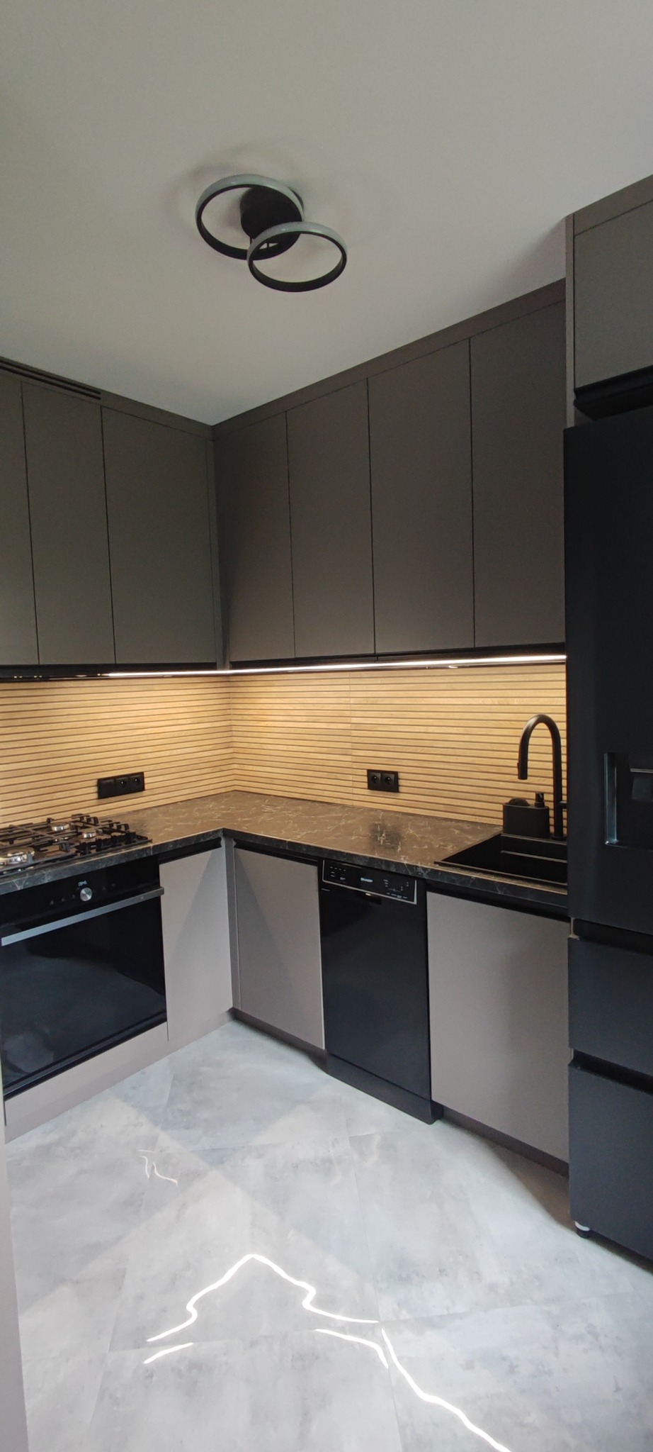 Nowoczesna kuchnia w odcieniach szarości z drewnianym panelem na ścianie, oświetleniem LED pod szafkami i marmurową podłogą. Minimalistyczny design i funkcjonalność.