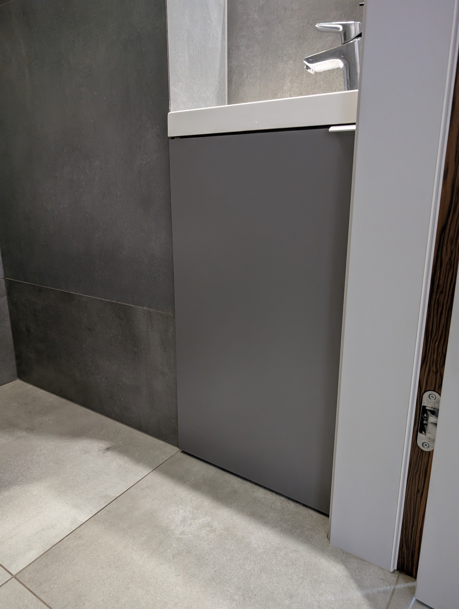 Szara szafka pod umywalką w nowoczesnej łazience z ciemnymi płytkami na ścianie i jasnymi na podłodze. Widoczny fragment baterii i drzwi.