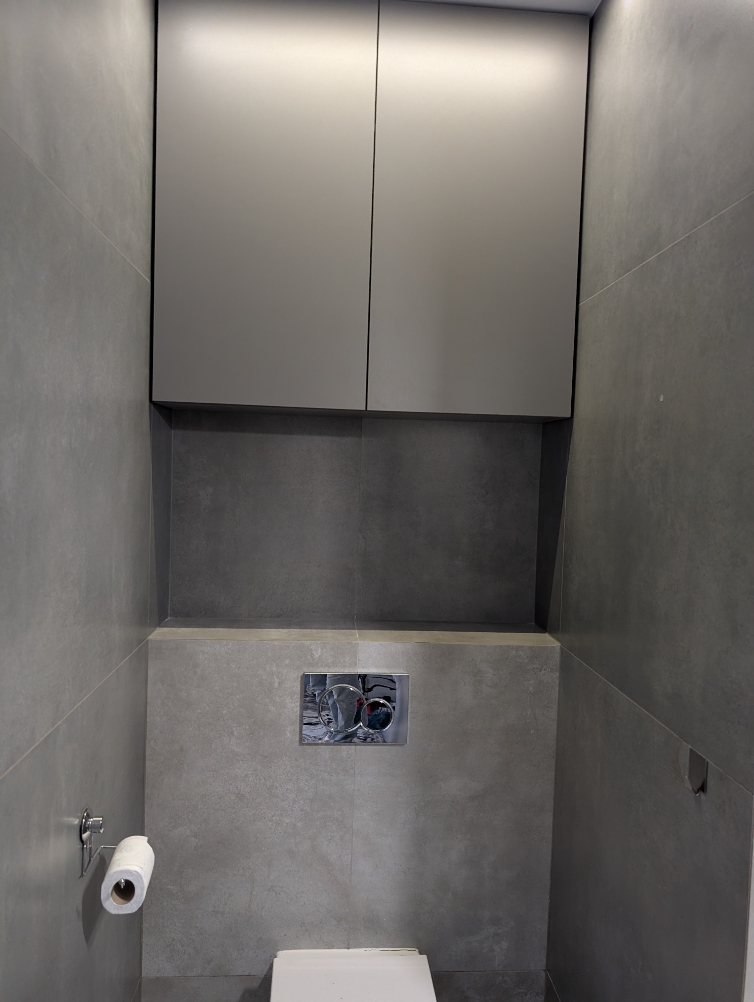 Nowoczesna toaleta w szarościach: wisząca szafka nad zabudowaną spłuczką, minimalistyczny design, płytki imitujące beton, uchwyt na papier toaletowy po lewej.