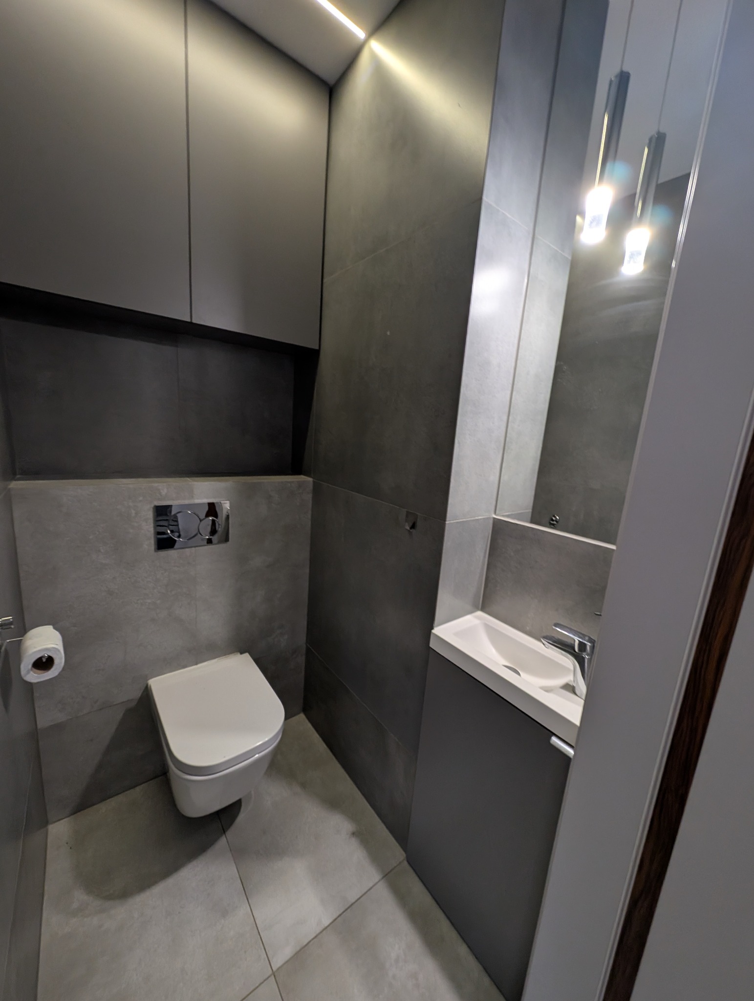 Nowoczesna, szara łazienka z podwieszaną toaletą, umywalką z szafką i lustrem z oświetleniem. Minimalistyczny design, płytki imitujące beton, eleganckie wykończenie.