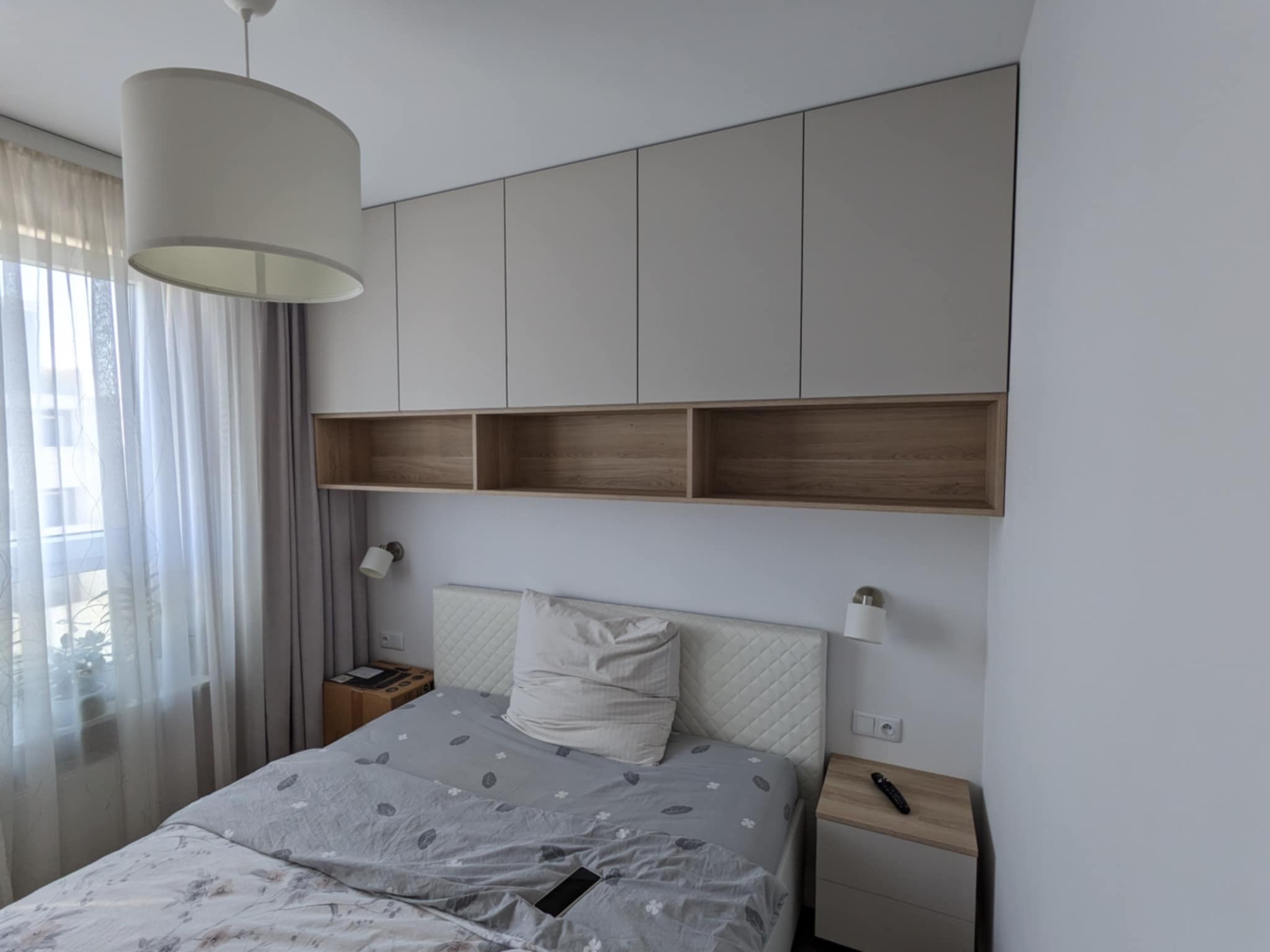 Jasna sypialnia z zabudową meblową nad łóżkiem: szafki z beżowymi frontami i otwartymi półkami z drewnianym dekorem. Minimalistyczny design, lampki nocne.