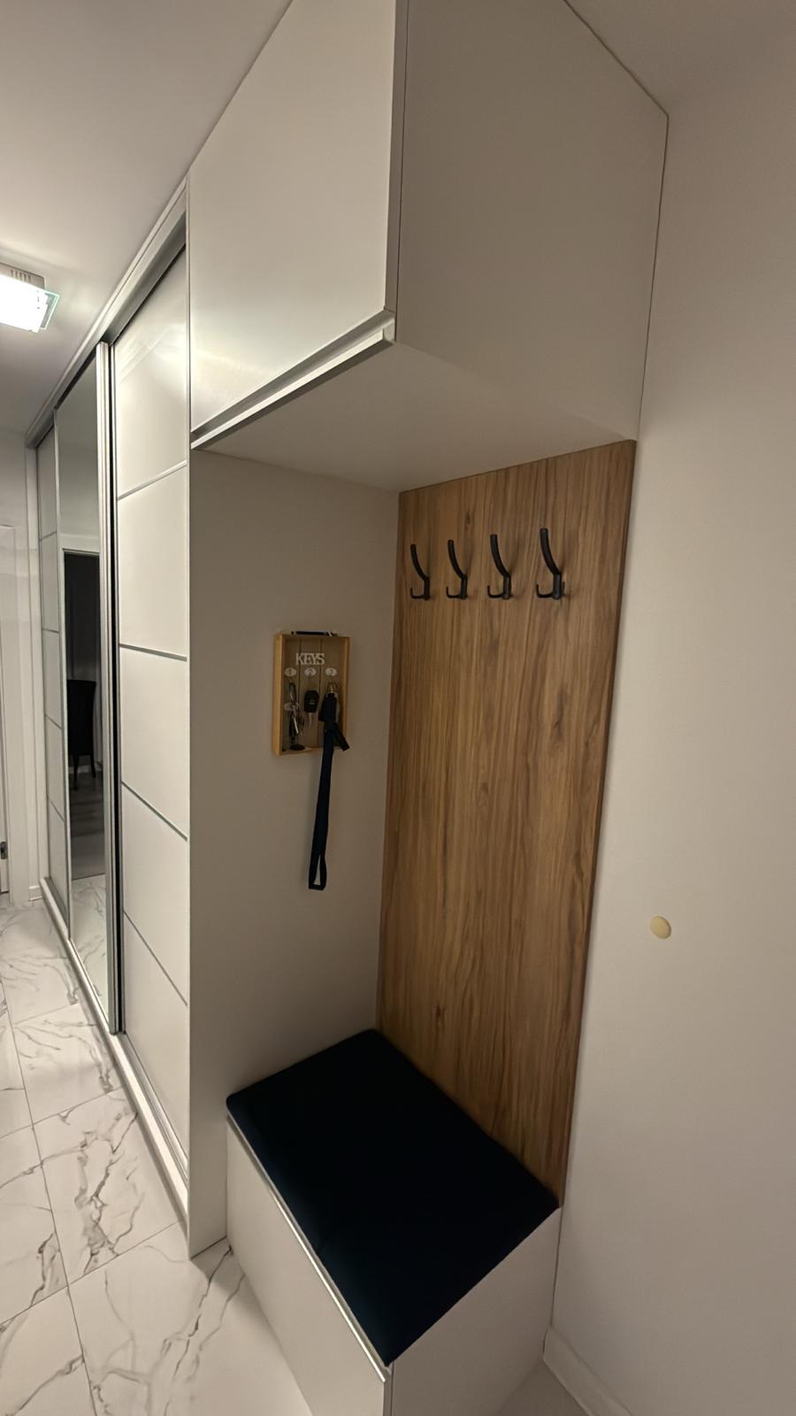Biała szafa przesuwna z lustrem i siedziskiem z wieszakami na ścianie. Widoczny organizer na klucze. Nowoczesny design, marmurowa podłoga.