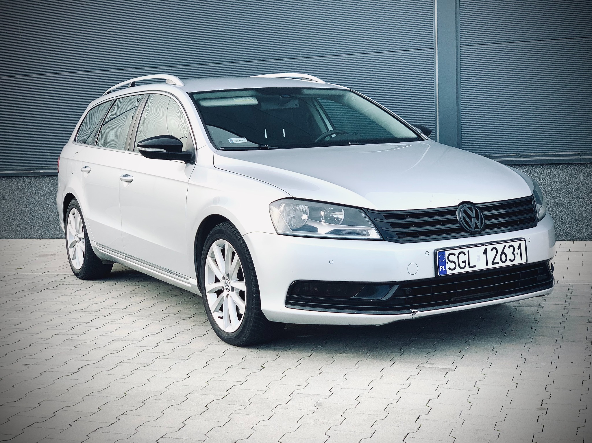 Srebrny Volkswagen Passat kombi, widok z przodu na tle szarej, żaluzjowej ściany. Auto zaparkowane na kostce brukowej. Tablica rejestracyjna widoczna.