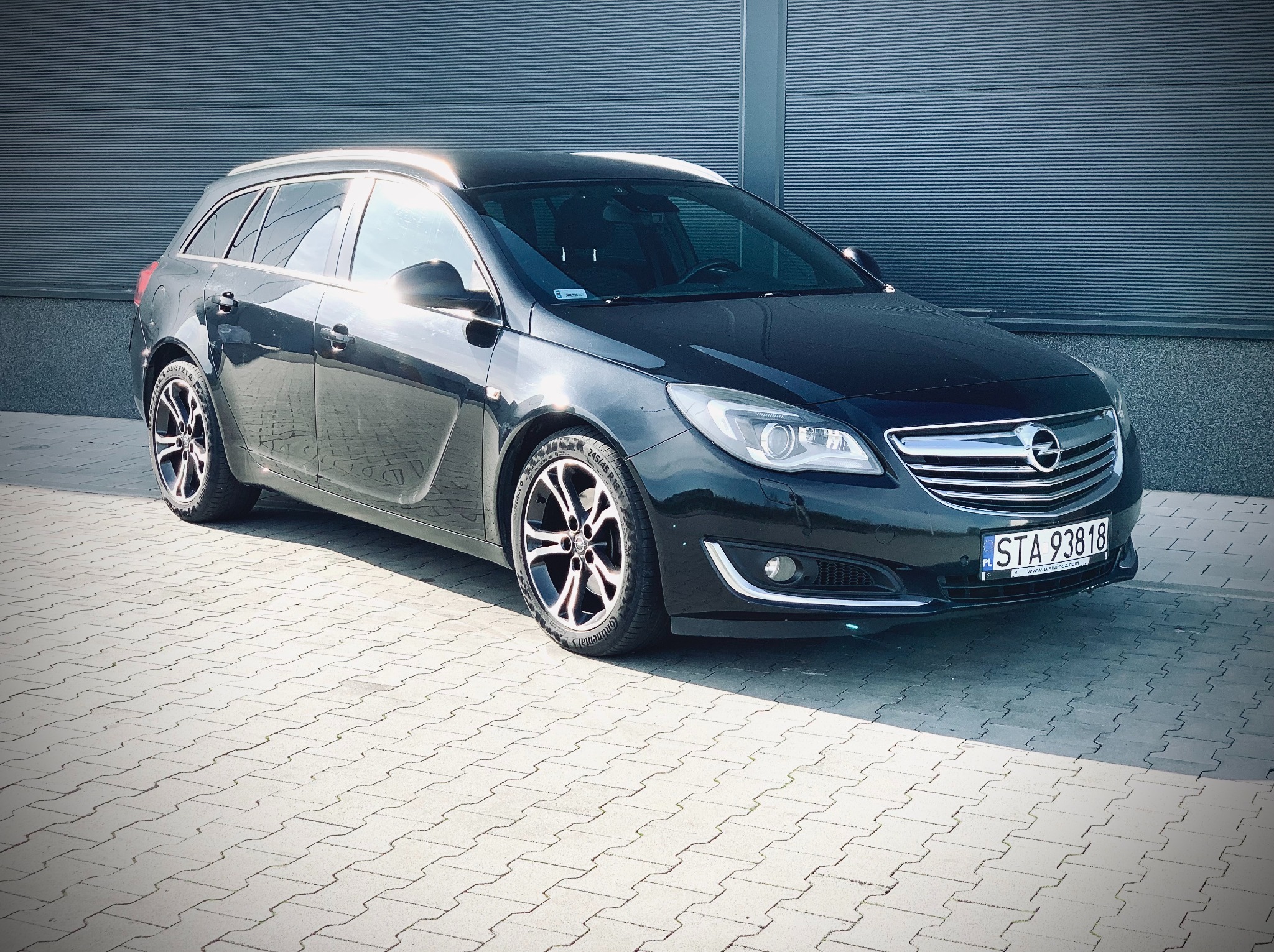 Czarna Opel Insignia kombi, zaparkowana przed metalową ścianą, z widoczną tablicą rejestracyjną STA 93818 i oponami Continental, prezentowana w jasnym świetle słonecznym.