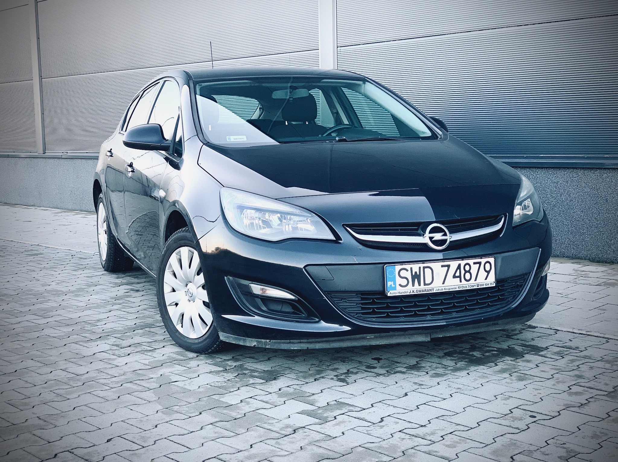 Czarny Opel Astra, widok z przodu na tle szarej, żłobkowanej ściany. Widoczna tablica rejestracyjna i felgi. Auto zaparkowane na kostce brukowej.