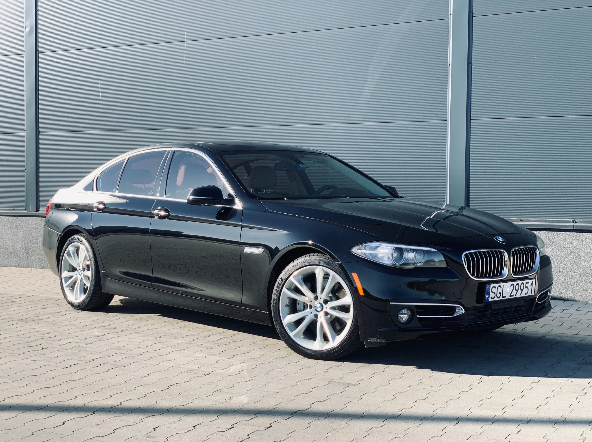 Czarna limuzyna BMW 535i, prezentowana na tle szarej ściany z poziomymi panelami. Auto zaparkowane na kostce brukowej, widoczne tablice rejestracyjne.
