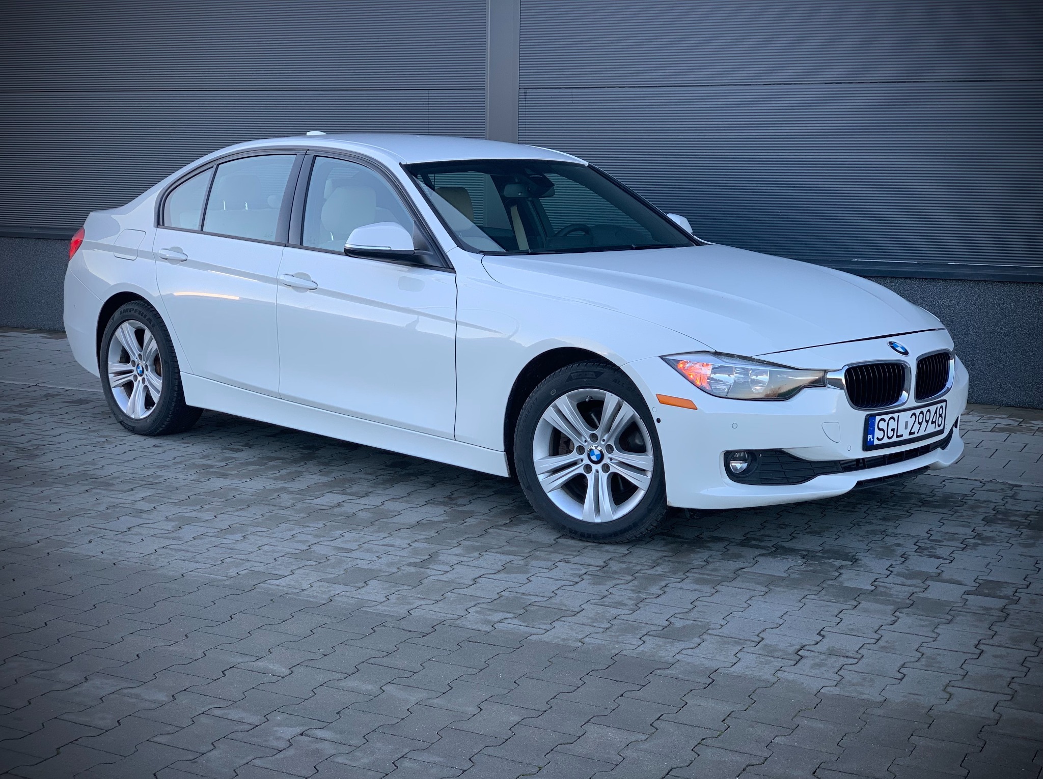 Białe BMW 328i, sedan, zaparkowane na szarym bruku przed budynkiem z poziomymi panelami. Widoczne tablice rejestracyjne SGL 29948 i felgi aluminiowe.