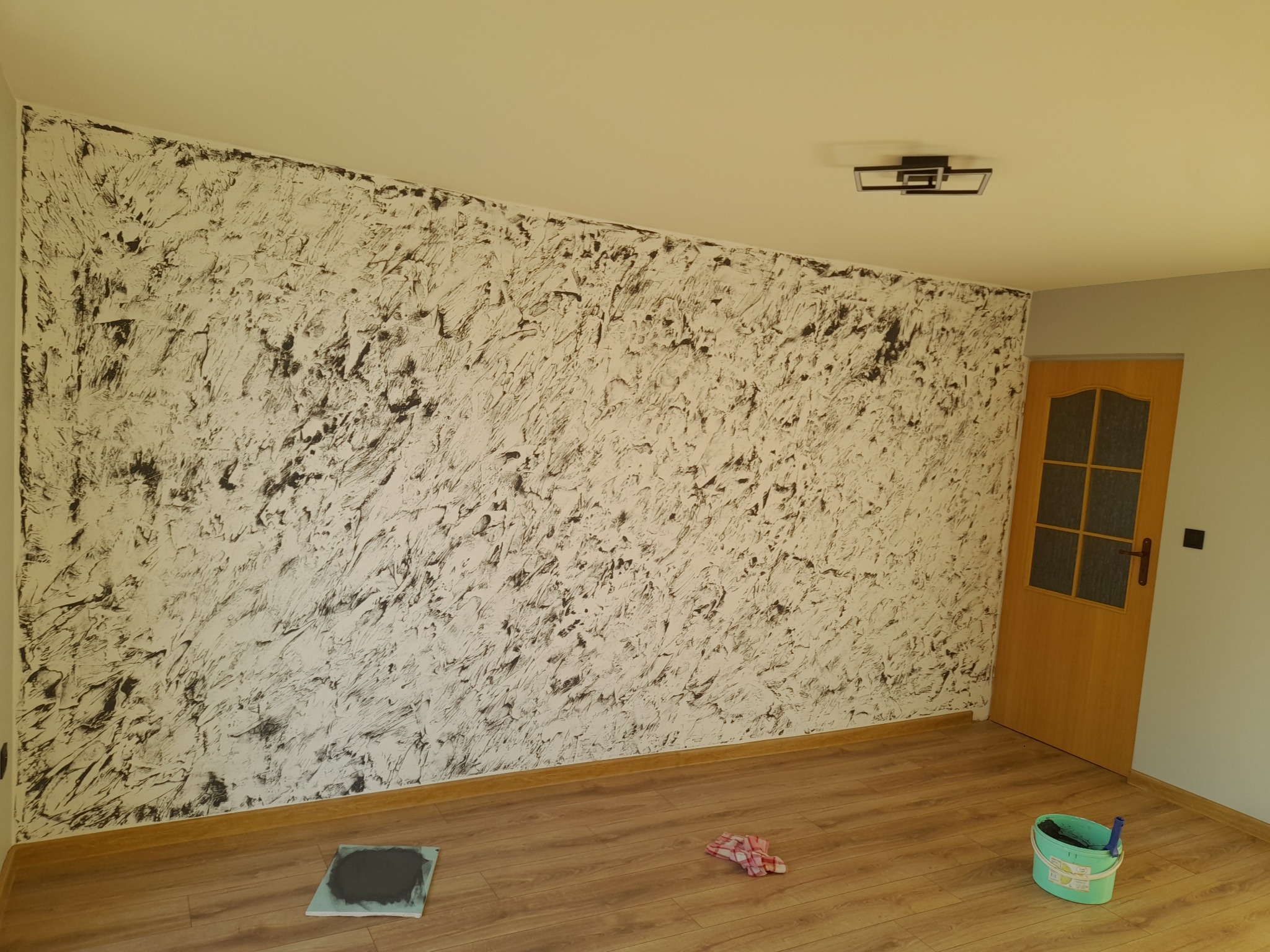 Ściana z efektem dekoracyjnym w odcieniach bieli i czerni. Na podłodze wiadro z farbą i szmatka. Widoczna drewniana podłoga i drzwi z szybkami.