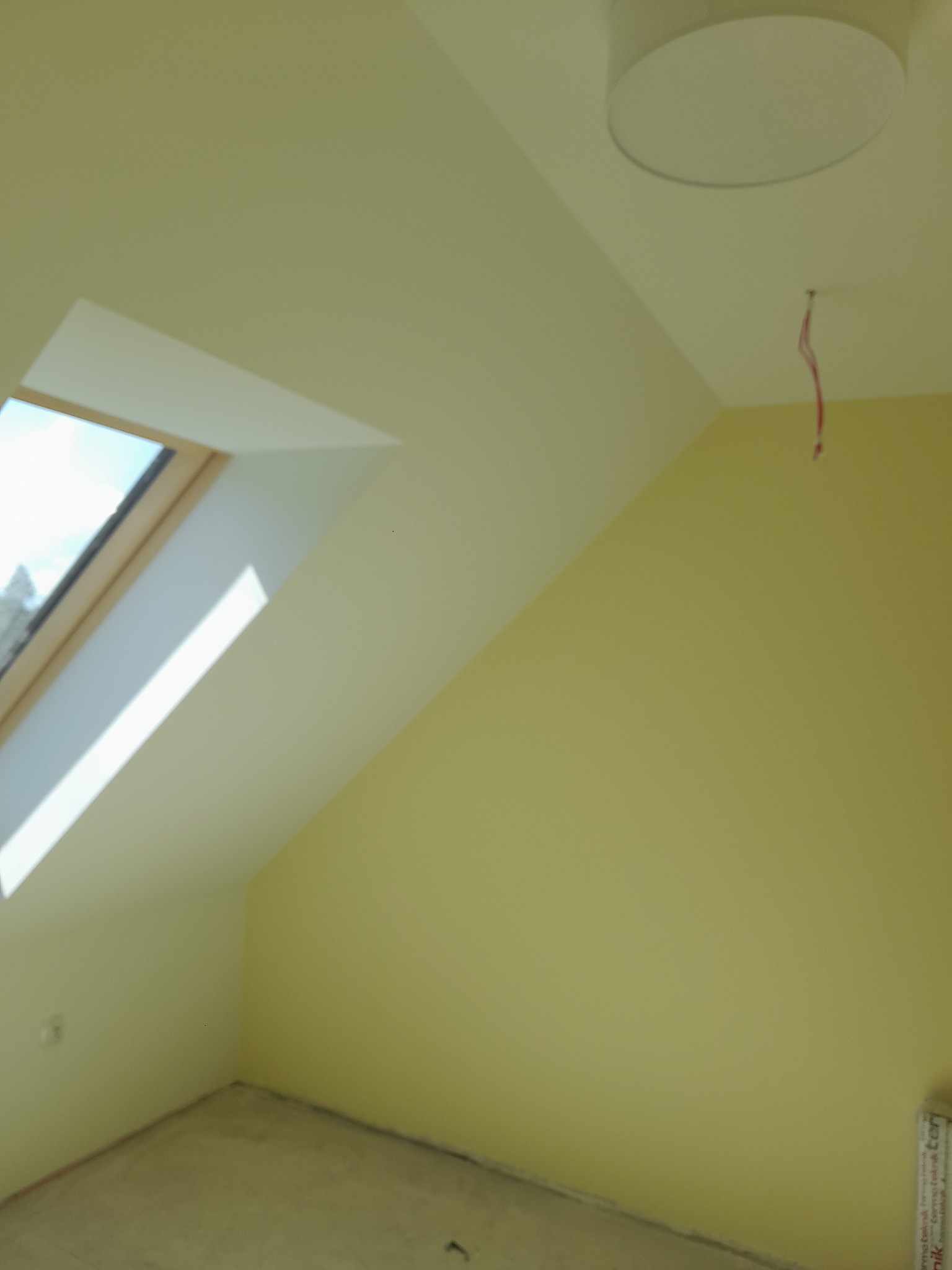 Pomalowane na jasnożółto poddasze z oknem dachowym i lampą sufitową. Widoczne ślady po przewodach elektrycznych. Ściany gładkie, podłoga przygotowana do dalszych prac.