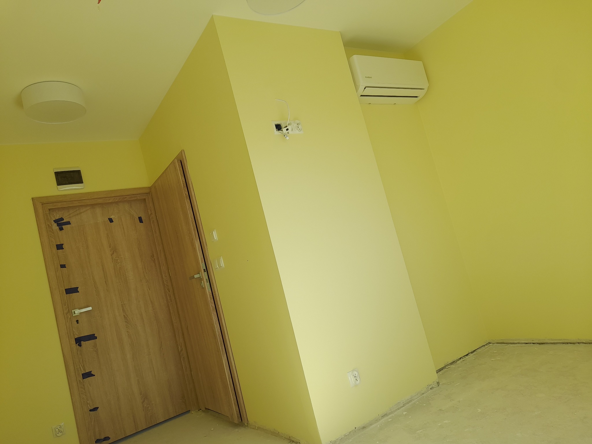 Świeżo pomalowane na żółto wnętrze z drewnianymi drzwiami zabezpieczonymi taśmą, klimatyzacją i gniazdkami elektrycznymi. W tle widoczny sufit z lampą.