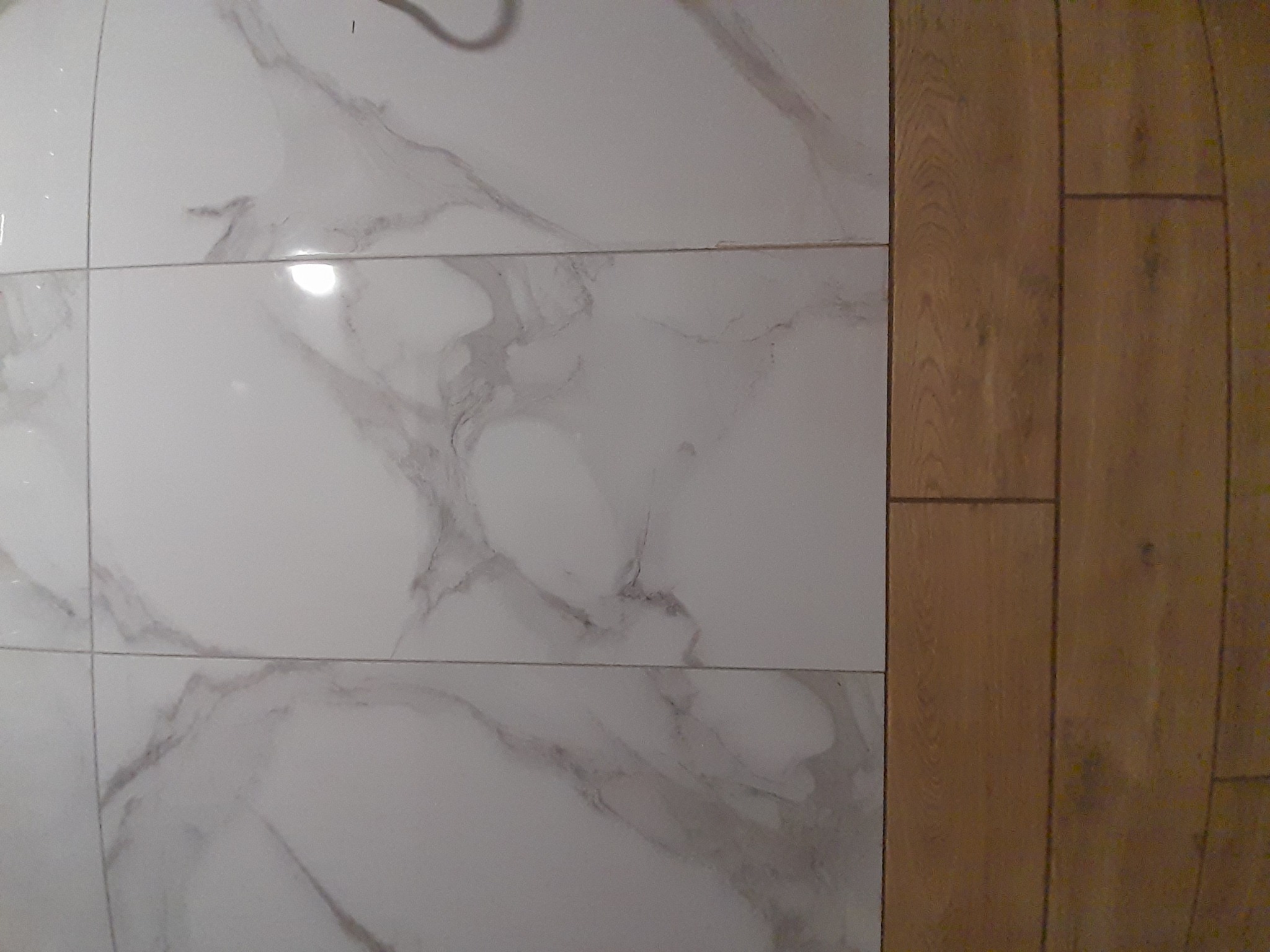 Połączenie podłogi drewnianej z marmurową, białą płytką w łazience. Widok na fragment ściany i podłogi, ukazujący precyzję wykonania i estetyczne wykończenie.