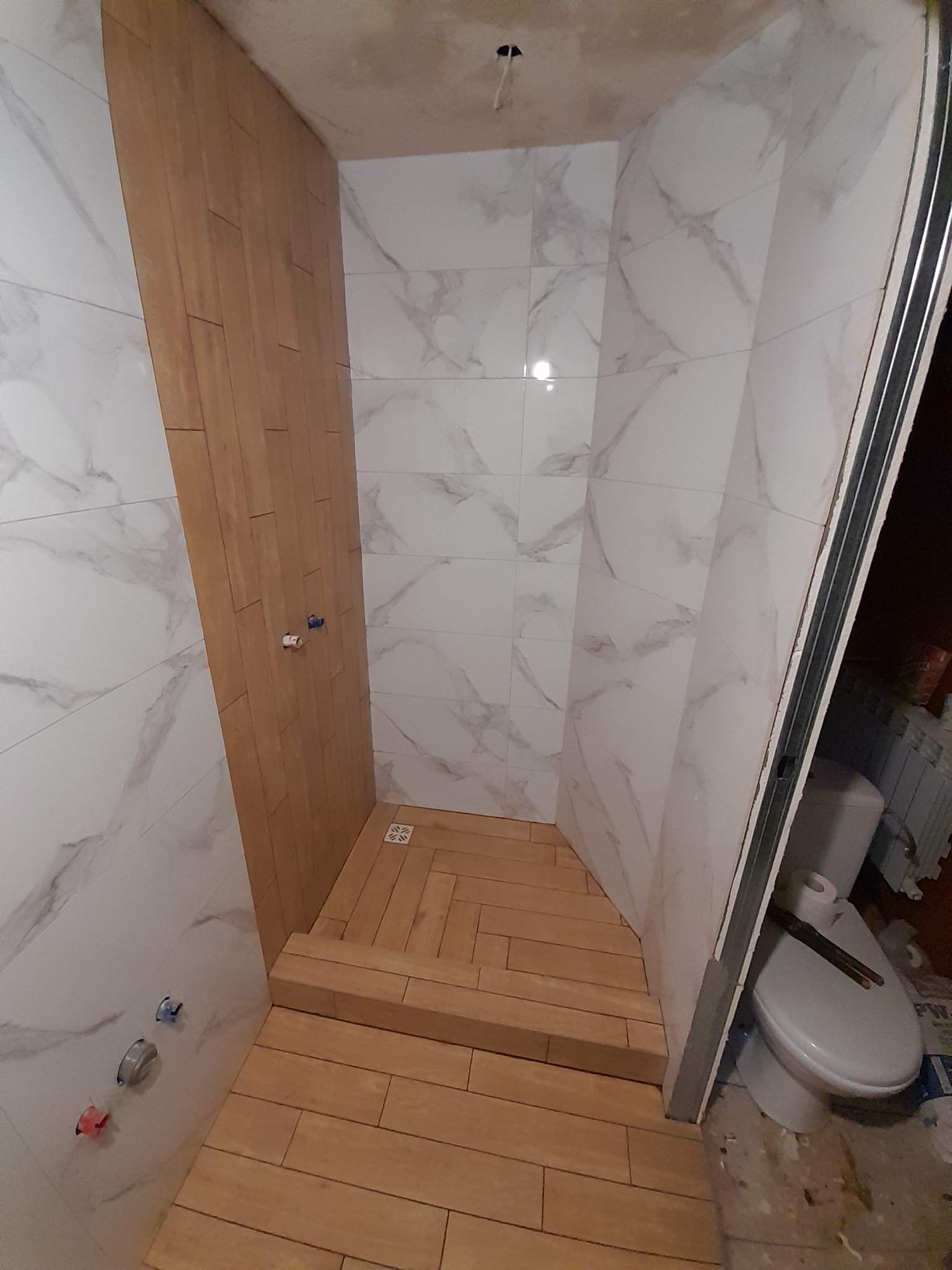 Wykończenie kabiny prysznicowej z brązowymi płytkami ułożonymi w jodełkę na podłodze i pionowo na ścianie, kontrastującymi z białymi płytkami imitującymi marmur.