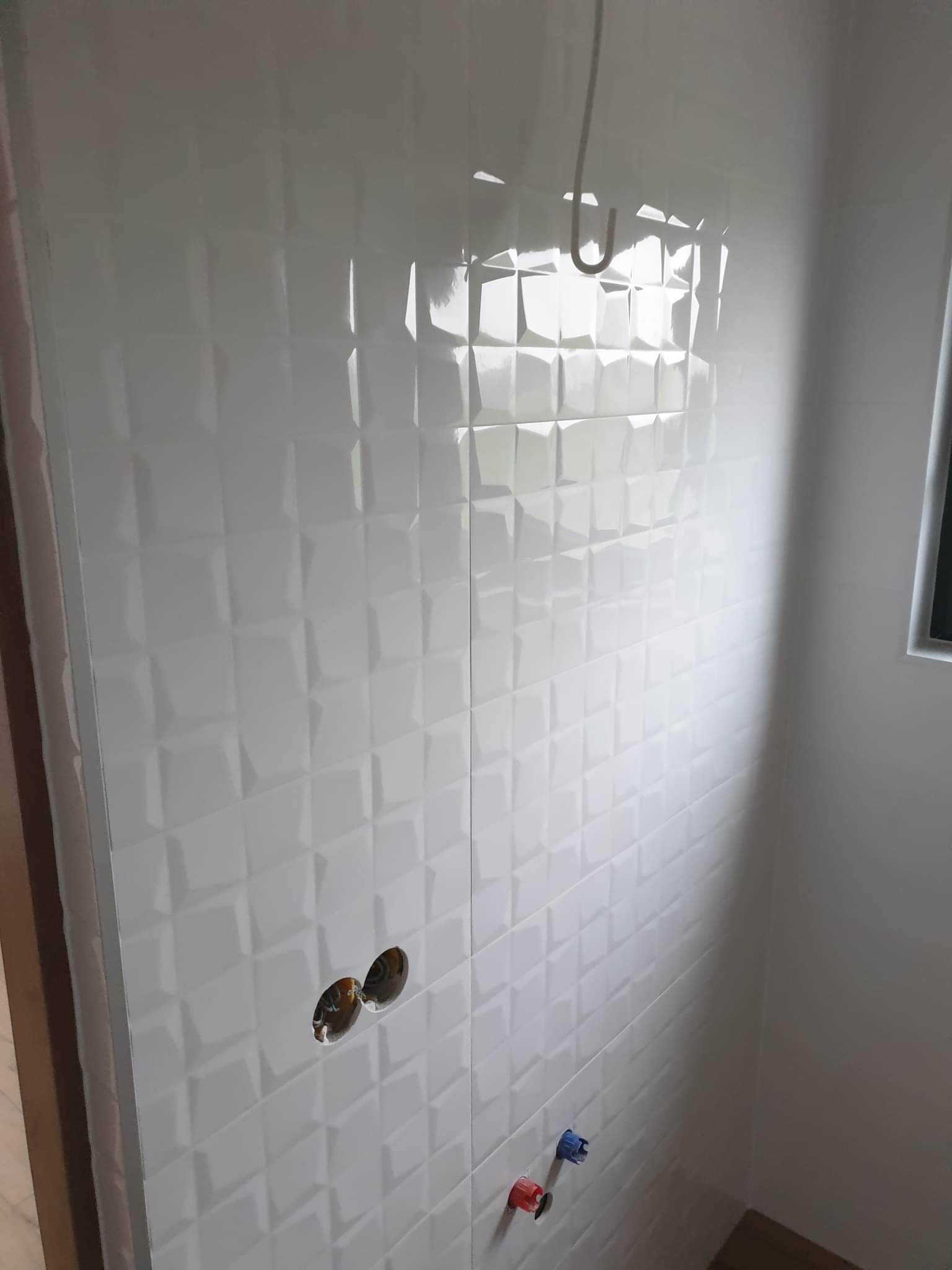 Ściana wyłożona białą, błyszczącą glazurą z geometrycznym wzorem. Widoczne otwory na baterie i rury doprowadzające wodę. Wykończenie łazienki w nowoczesnym stylu.