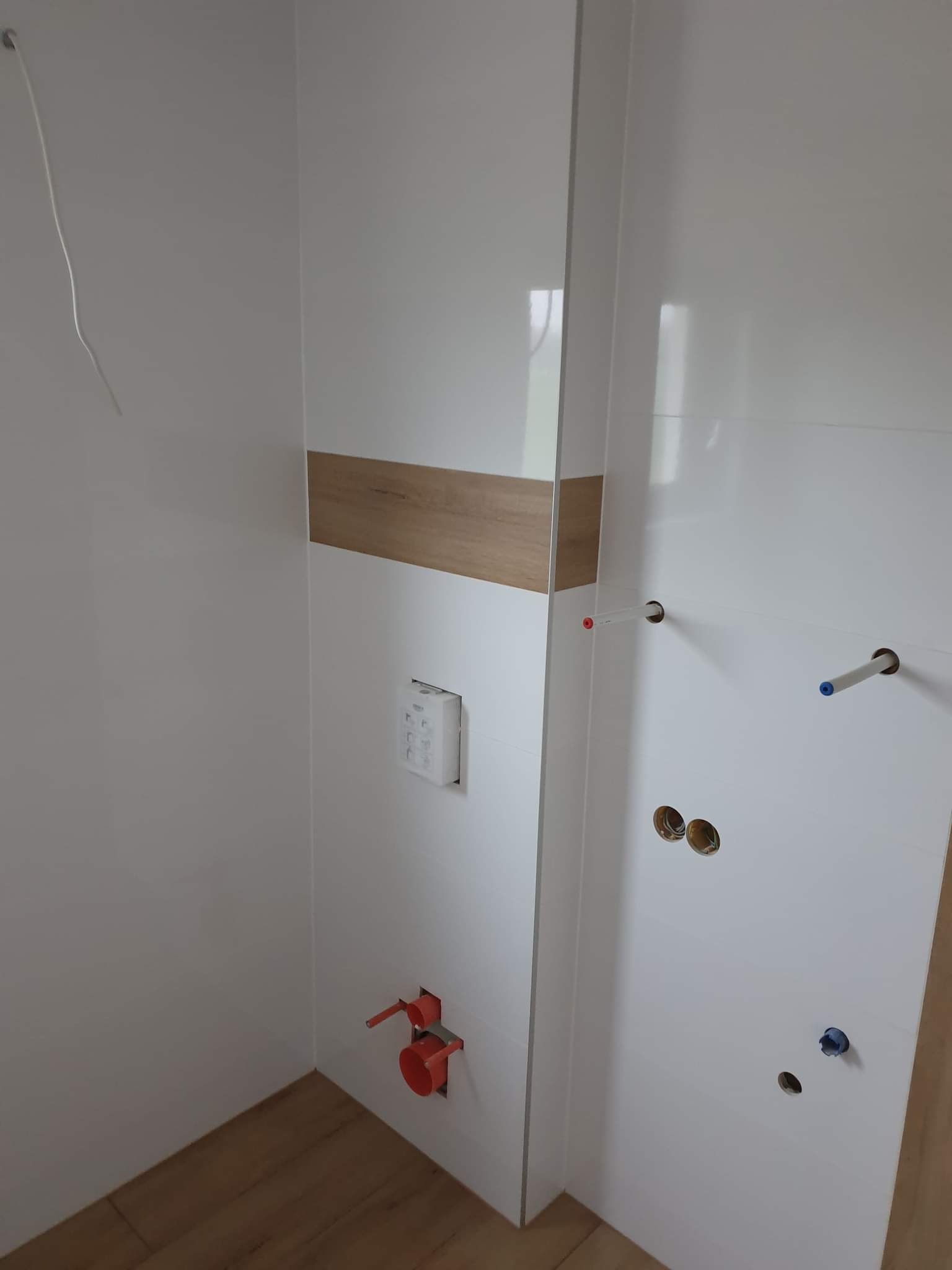 Białe, błyszczące płytki w łazience z drewnianym akcentem, widoczne przyłącza wodne i kanalizacyjne. Częściowo ułożona podłoga z paneli.