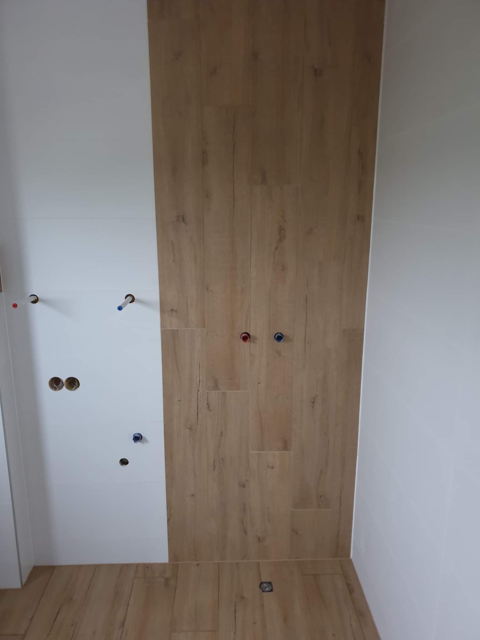 Wykończona ściana i podłoga w łazience z drewnopodobnych płytek. Widoczne wyprowadzenia wodne. Minimalistyczny design, jasne kolory.