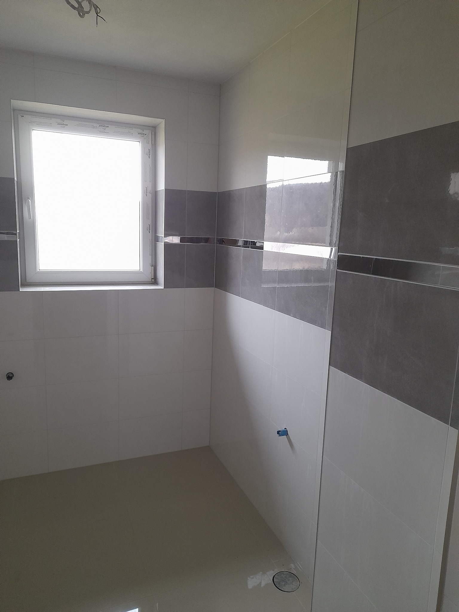 Wykończona łazienka z biało-szarymi płytkami, okno z białymi ramami. Widoczne fragmenty instalacji. Minimalistyczny design wnętrza, jasne oświetlenie.