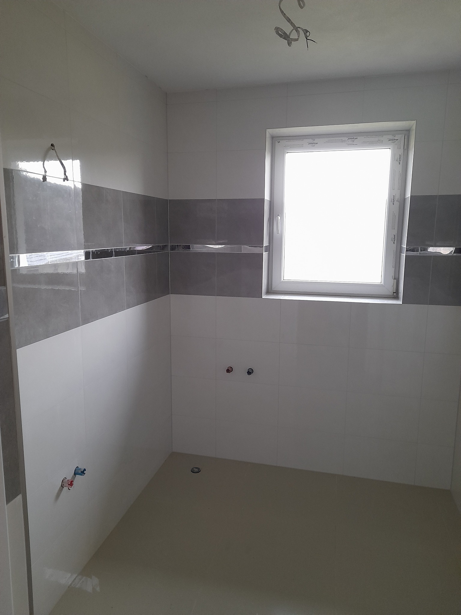 Wykończona łazienka z szarym pasem płytek, białe ściany, okno z białym obramowaniem, widoczne przyłącza wodne i odpływ podłogowy. Minimalistyczny design.