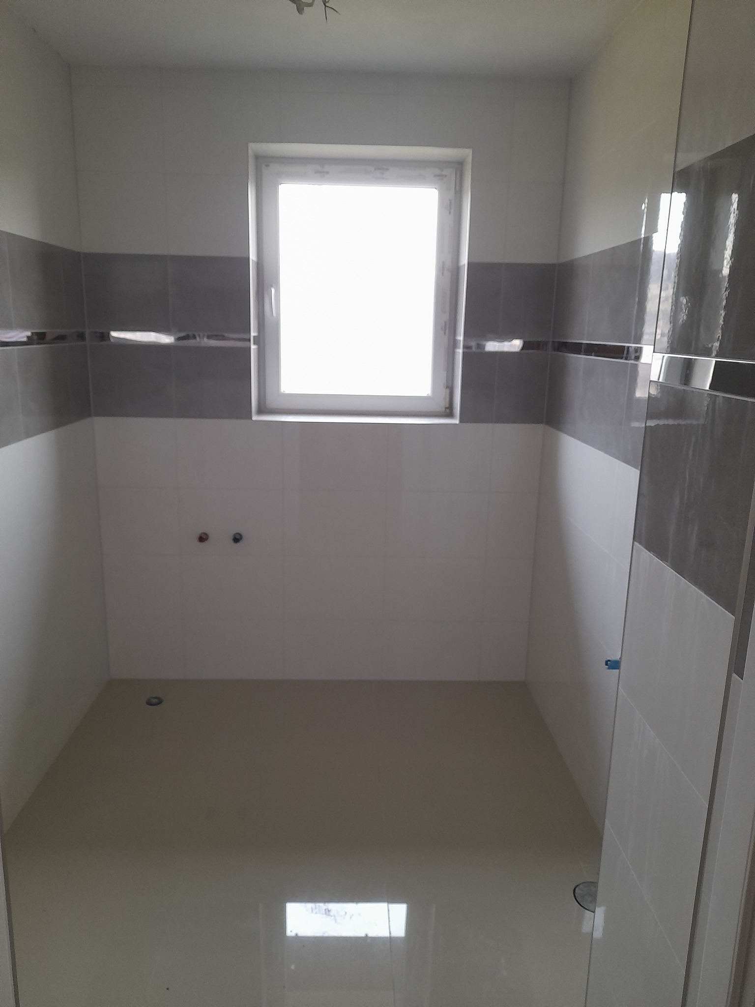 Wykończona łazienka z jasnymi płytkami i szarym pasem dekoracyjnym. Widoczne przyłącza wodne i odpływ. Okno w centralnej części ściany.