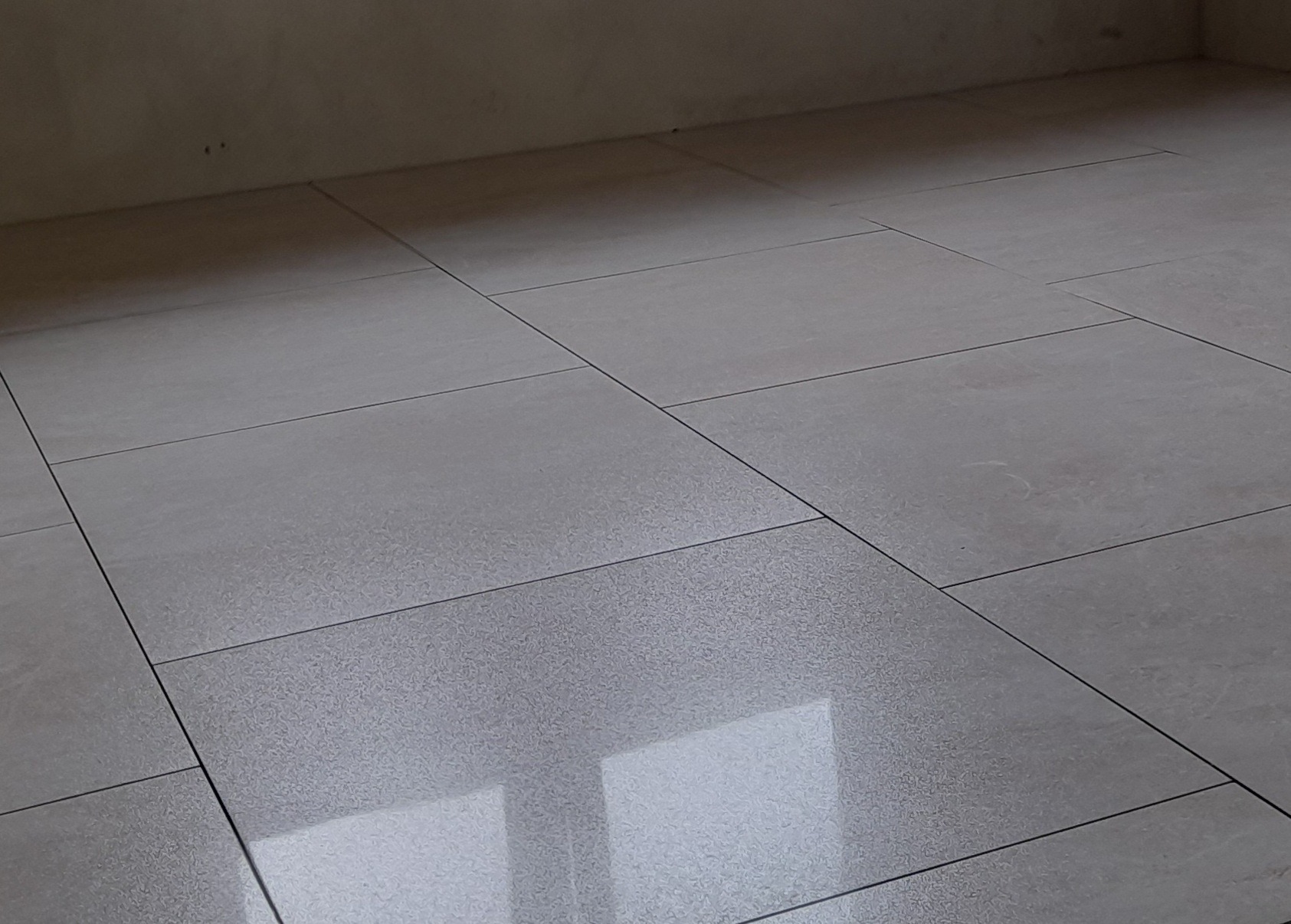 Jasnoszare, duże płytki podłogowe z ciemniejszą fugą, ułożone w regularny wzór. Widoczna ściana w tle i odbicie okna na powierzchni płytek.